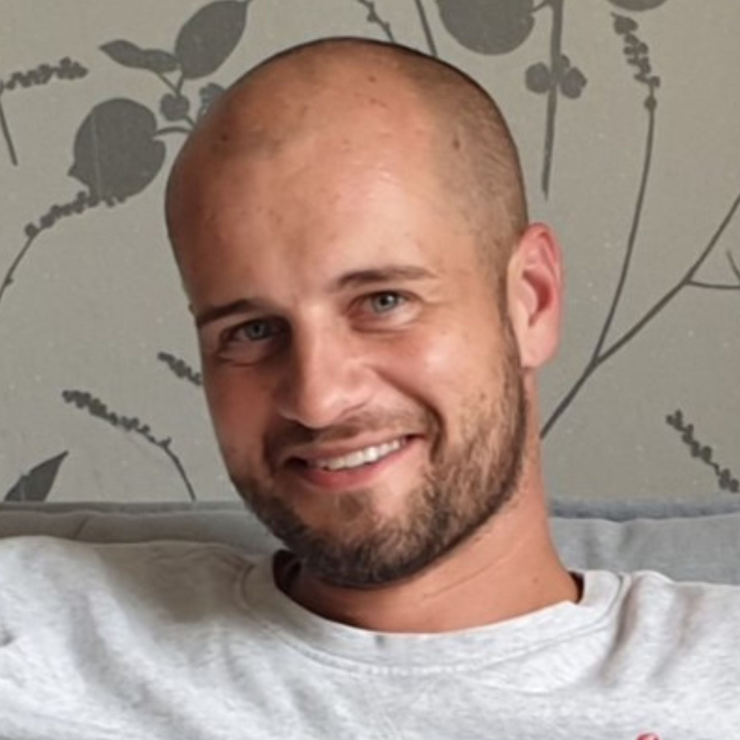 A man with a shaved head looking towards the camera smiling. The photo has been taken a getting a patterned background and the man is wearing a white/grey top.