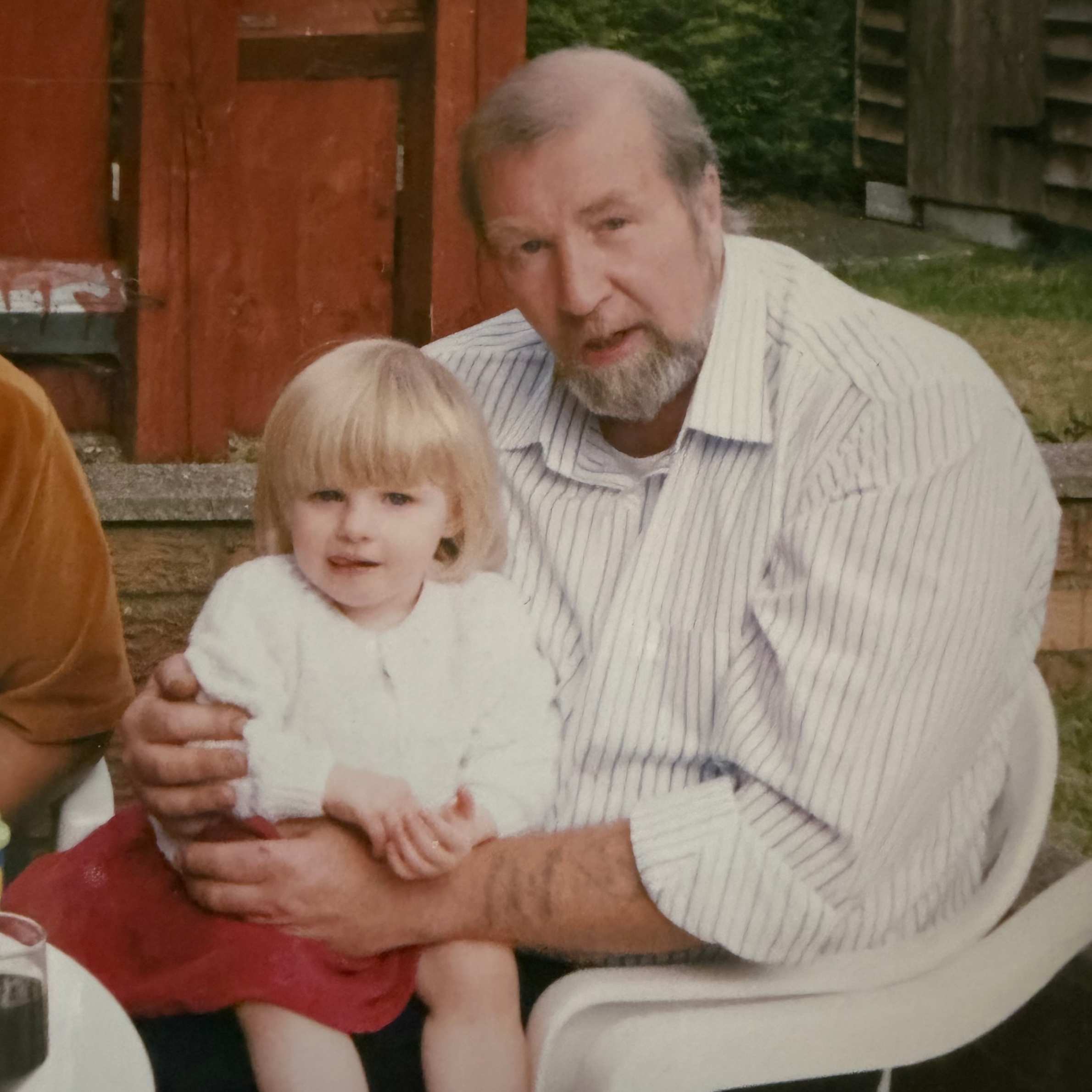 A photo of a grandparent with their grandchild sat in a plastic garden chair. The man is wearing a white striped shirt and the little girl has a white cardigan and red dress. They are both sat outside in the garden