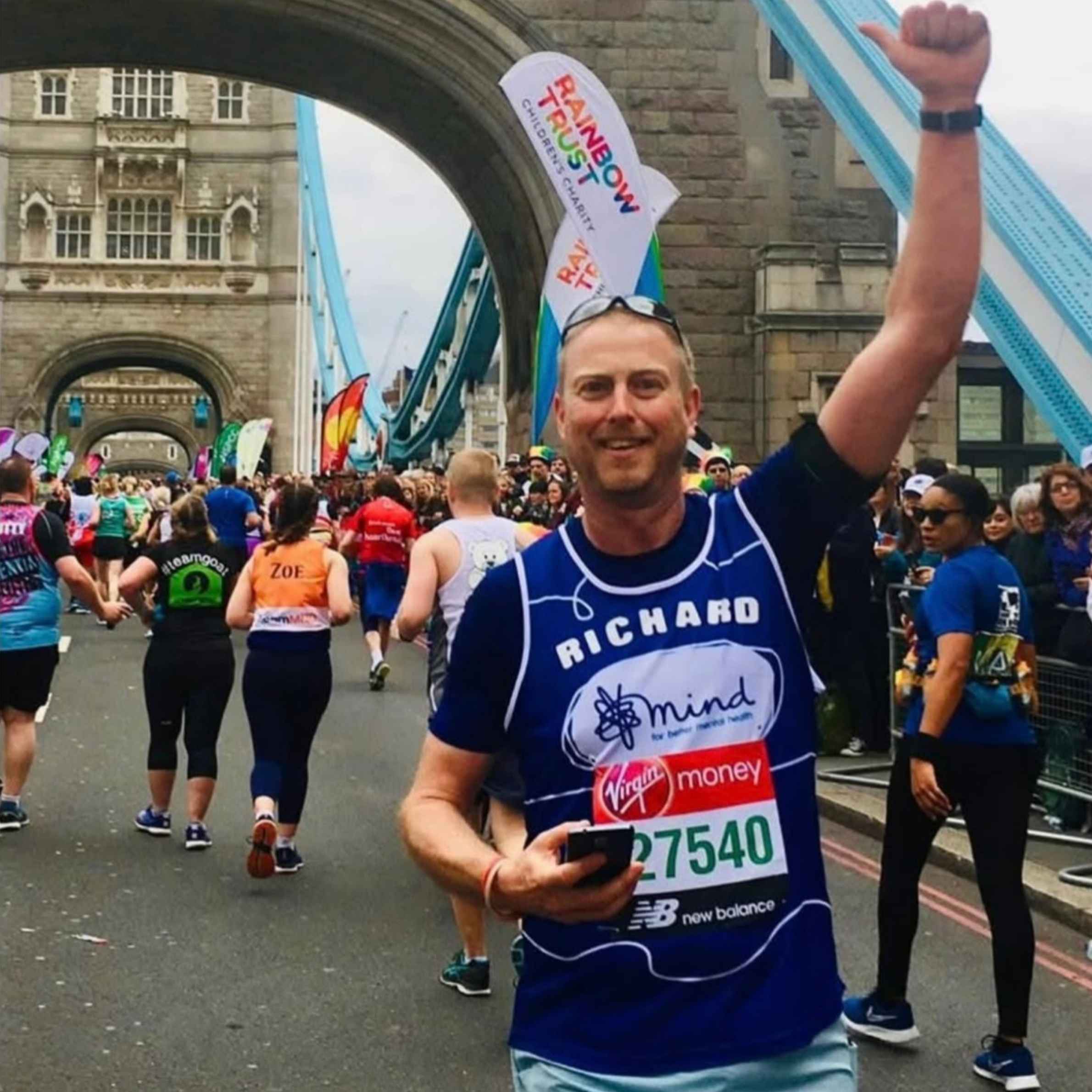 A Man on London Bridge running a marathon for Mind Charity. He is wearing a dark top and blue shorts holding one arm in the air
