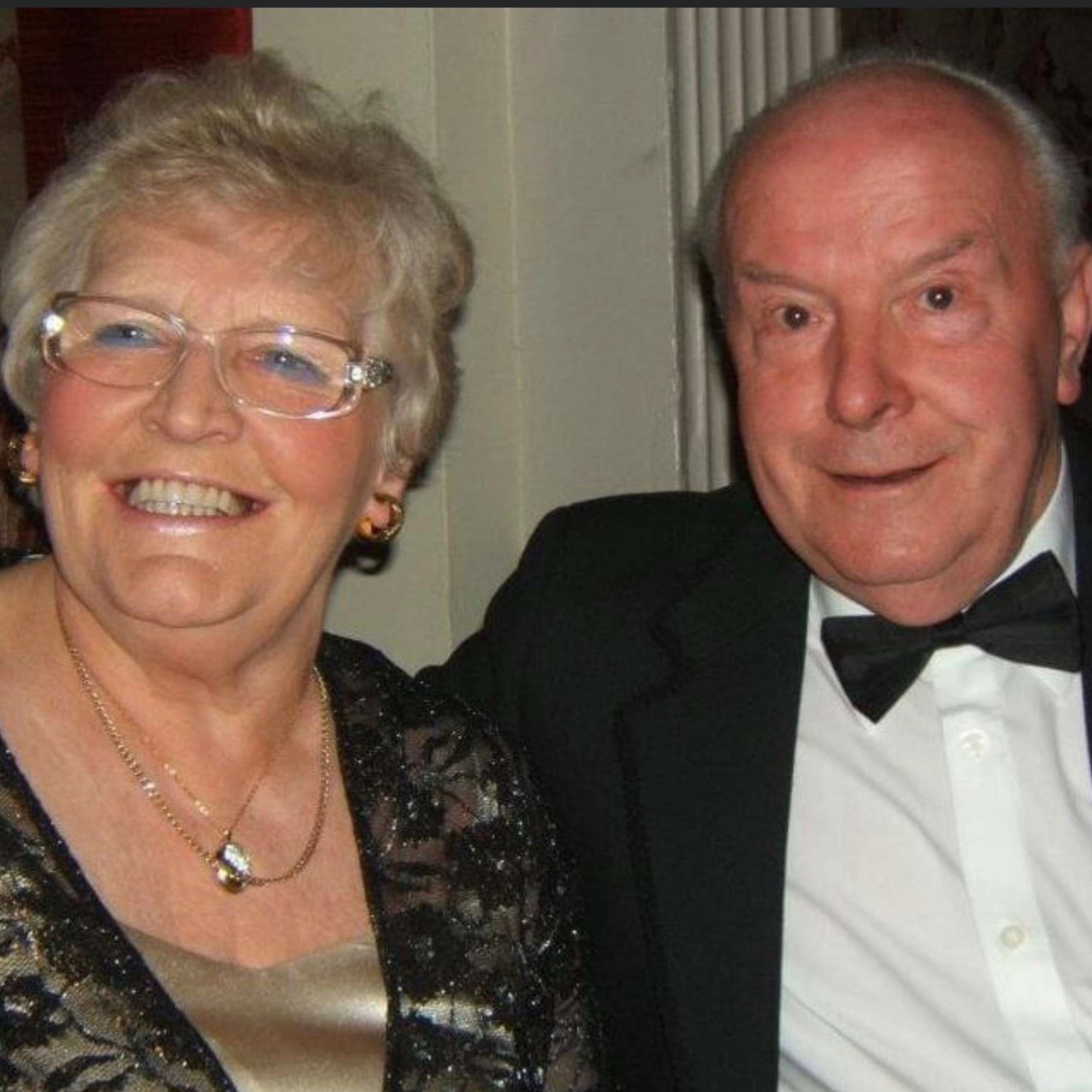 A set of grandparents smiling at the camera. They are wearing formal clothing so the man is wearing a black tie suit and the woman is wearing a black and gold top. They are sat against a plain background.