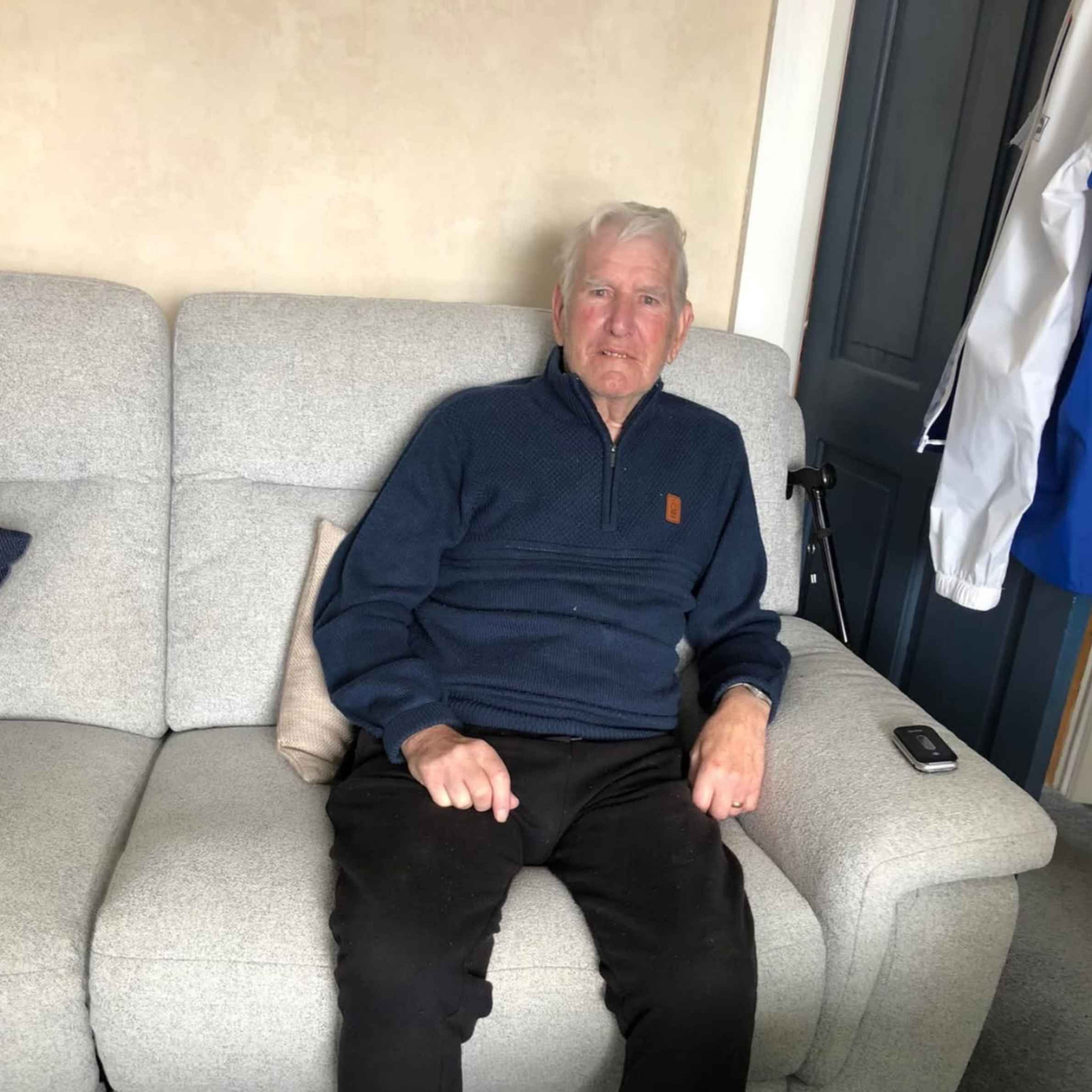 A man sat on a light grey sofa in a room with beige walls and a black door. The man is wearing a navy jumper with black trousers, sat in a relaxed upright position. 
