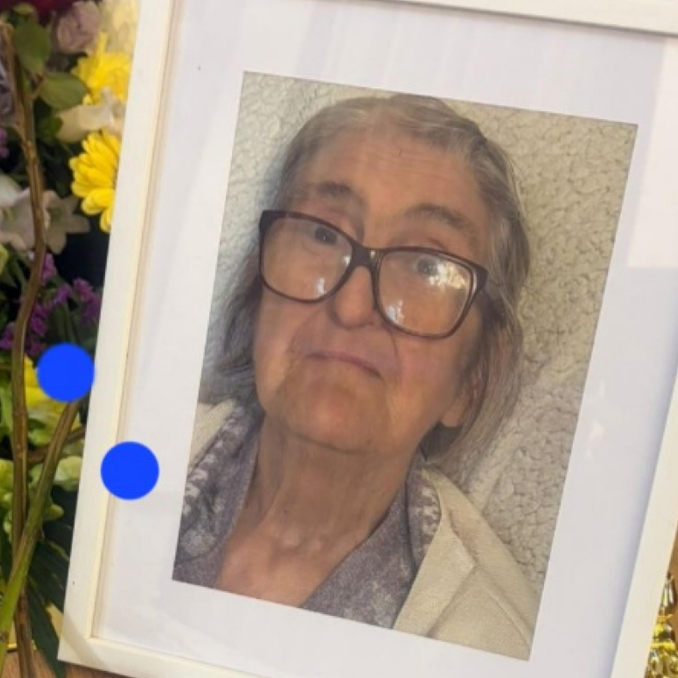 photo of mother in white frame. She was wearing glasses and a blue top with a cream cardigan. There are flowers in the background