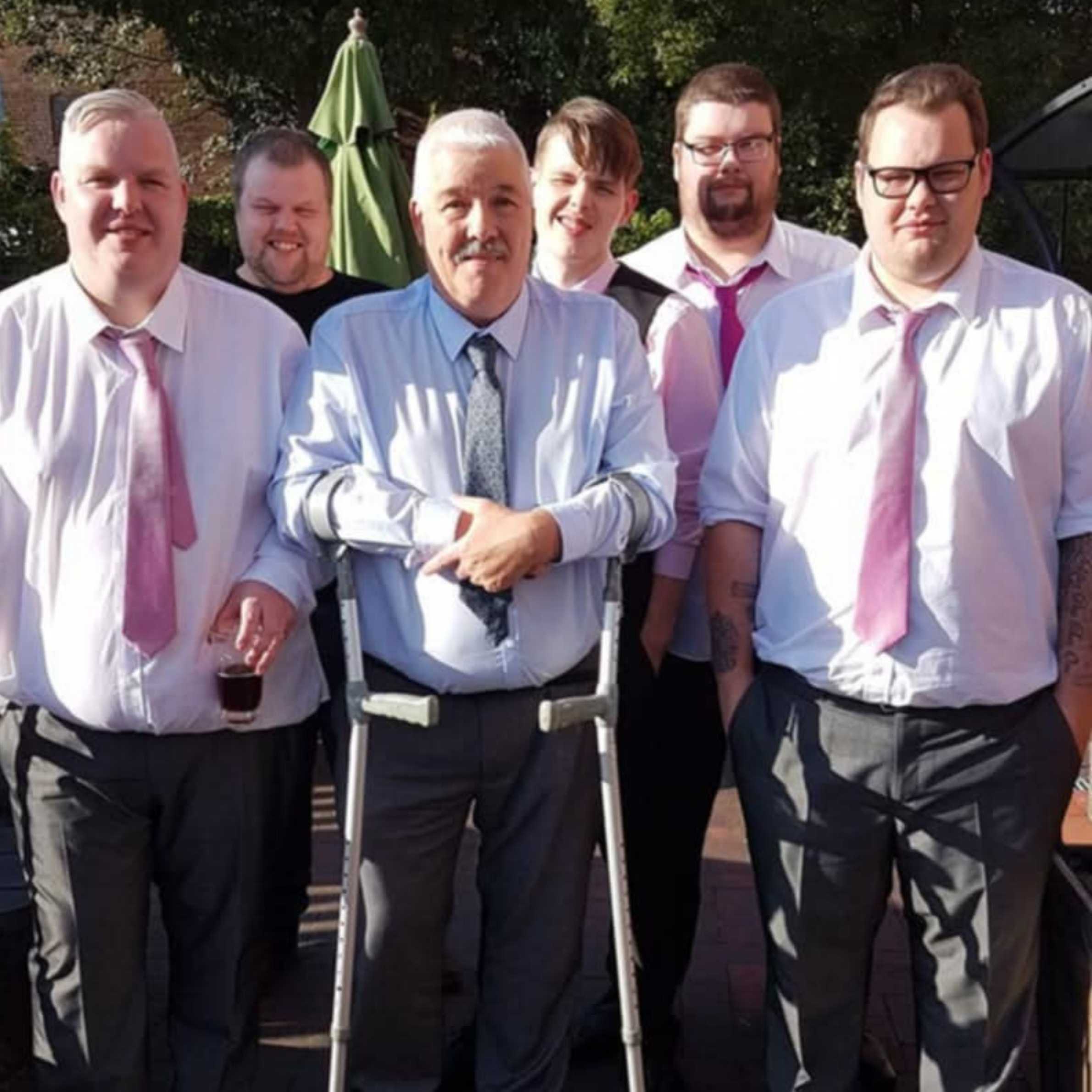 six men in the photo, three of which are wearing pink ties and white shirts. They are gathered around an older man with crutches. He's wearing a white shirt with a grey tie and they are all smiling looking towards the camera.