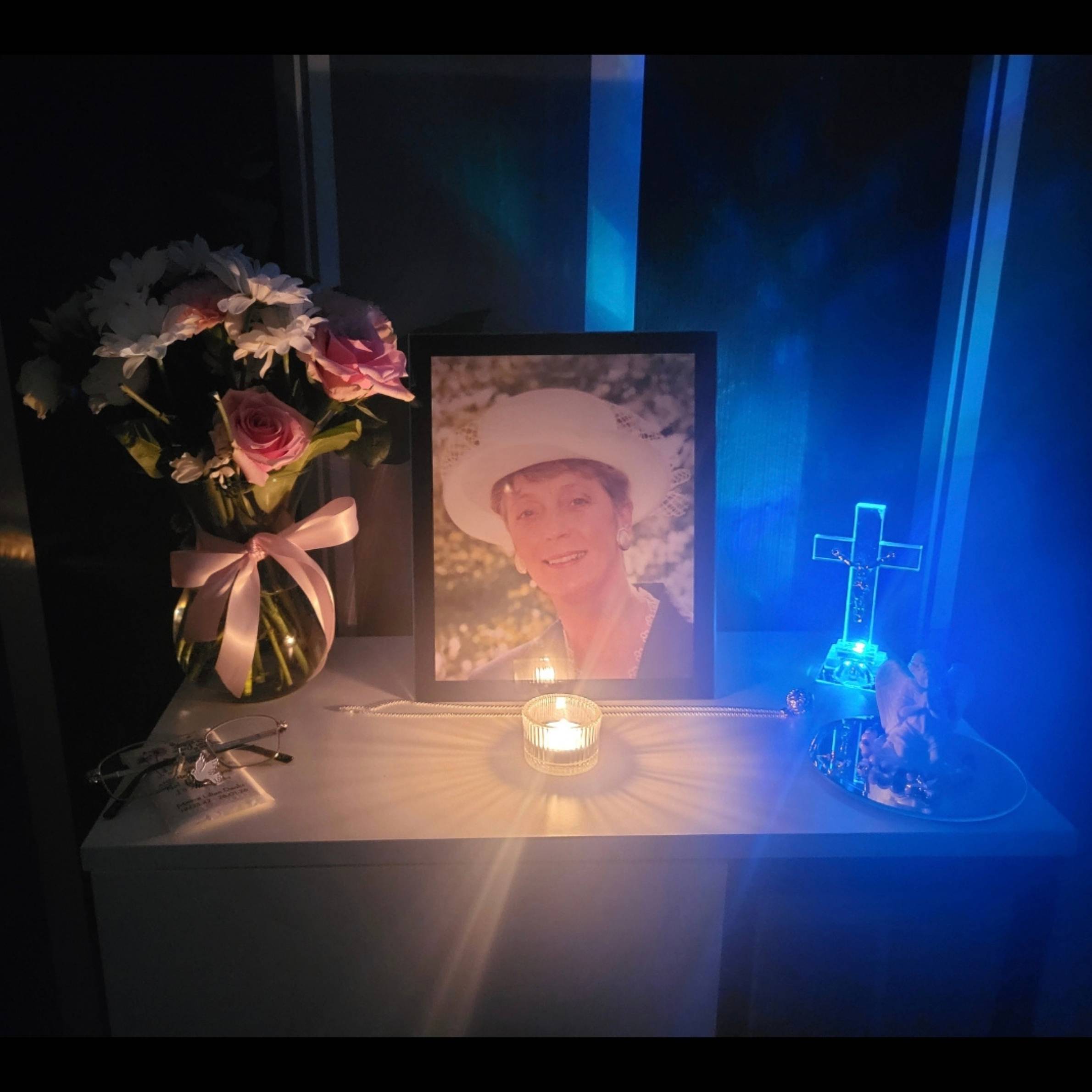 a photo of a loved one who has passed away on a table in a black frame with a lit candle in front of the frame. There is a bunch of white flowers with pink roses in a vase to the left of the frame and a blue light up cross to the right.