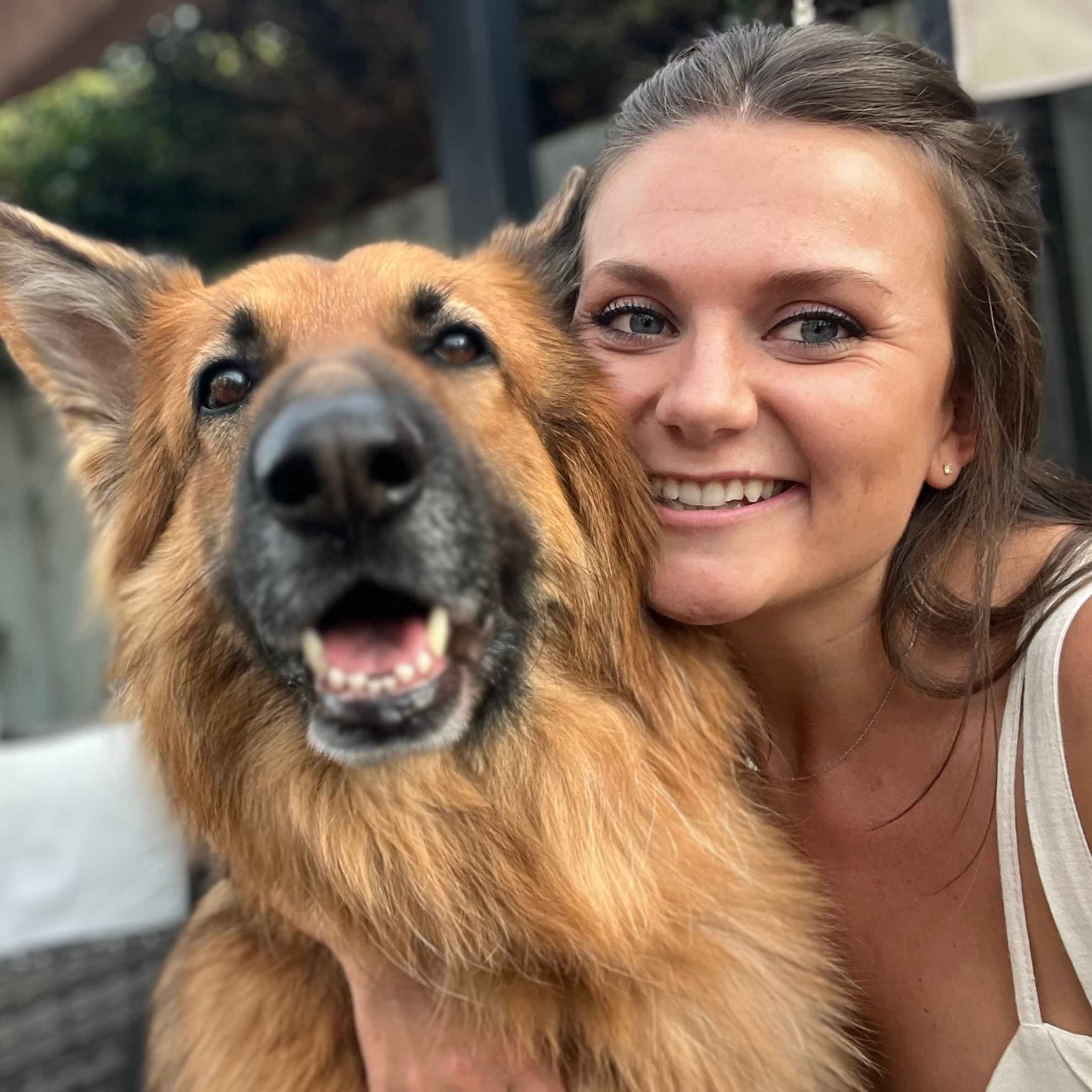brown German Shepherd to the left of the photo with black nose. To the right is a lady smiling wearing a cream strappy top, she has brown hair.