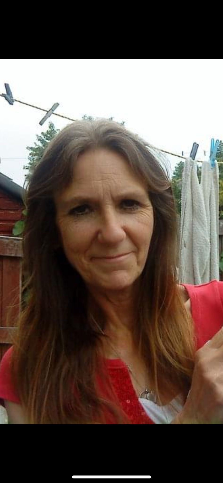 Lady with long brown hair smiling with closed mouth. shes wearing a red top and stood in front of a washing line and brown shed