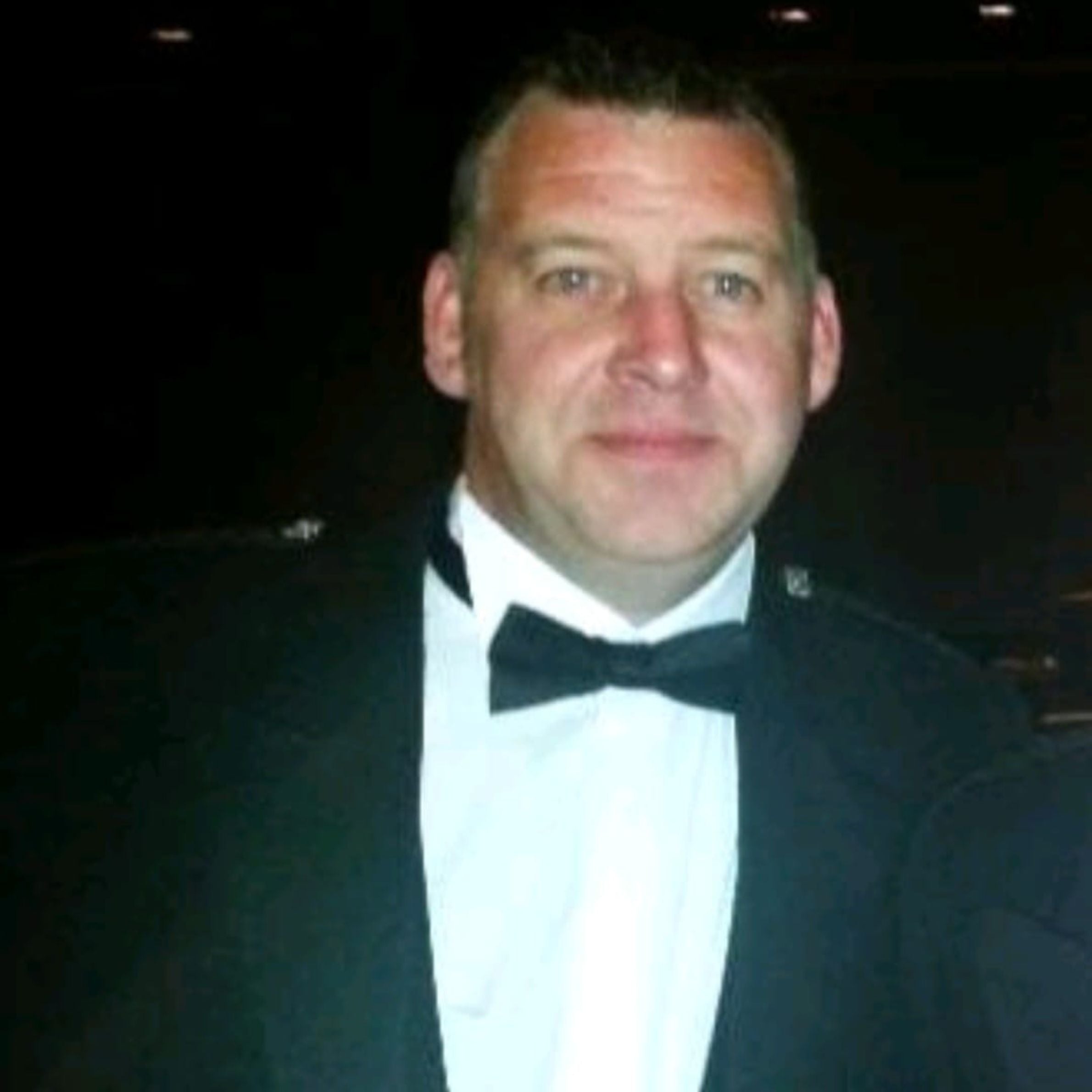 man wearing a black suit with a black bow. he has short dark hair 