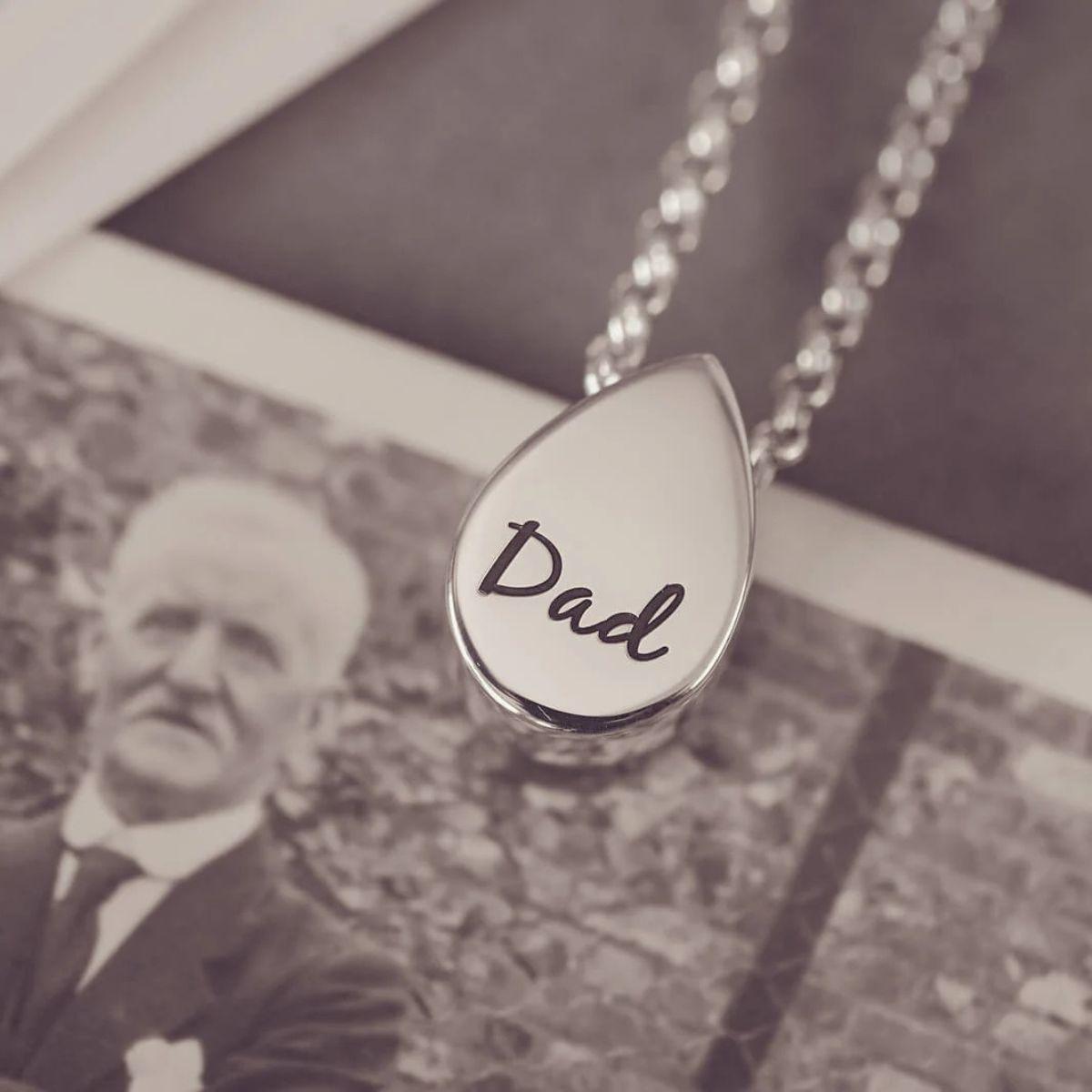 Teardrop-shaped pendant with 'Dad' engraved on it, displayed over a black and white photo of an older man.