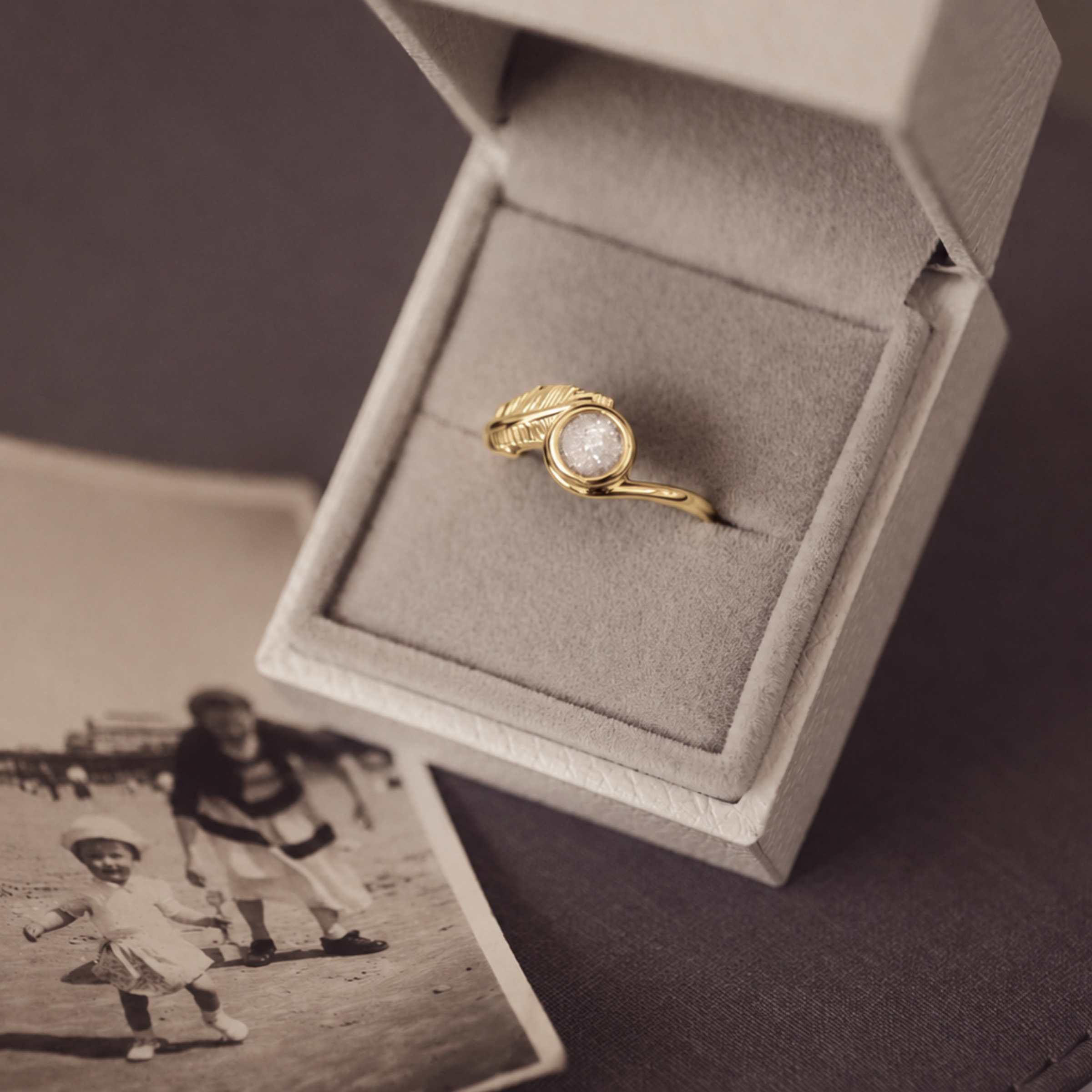 A gold ring with a textured feather detail and a round center stone, displayed inside an open light jewelry box. A vintage-style black-and-white photo lies nearby on a dark surface.