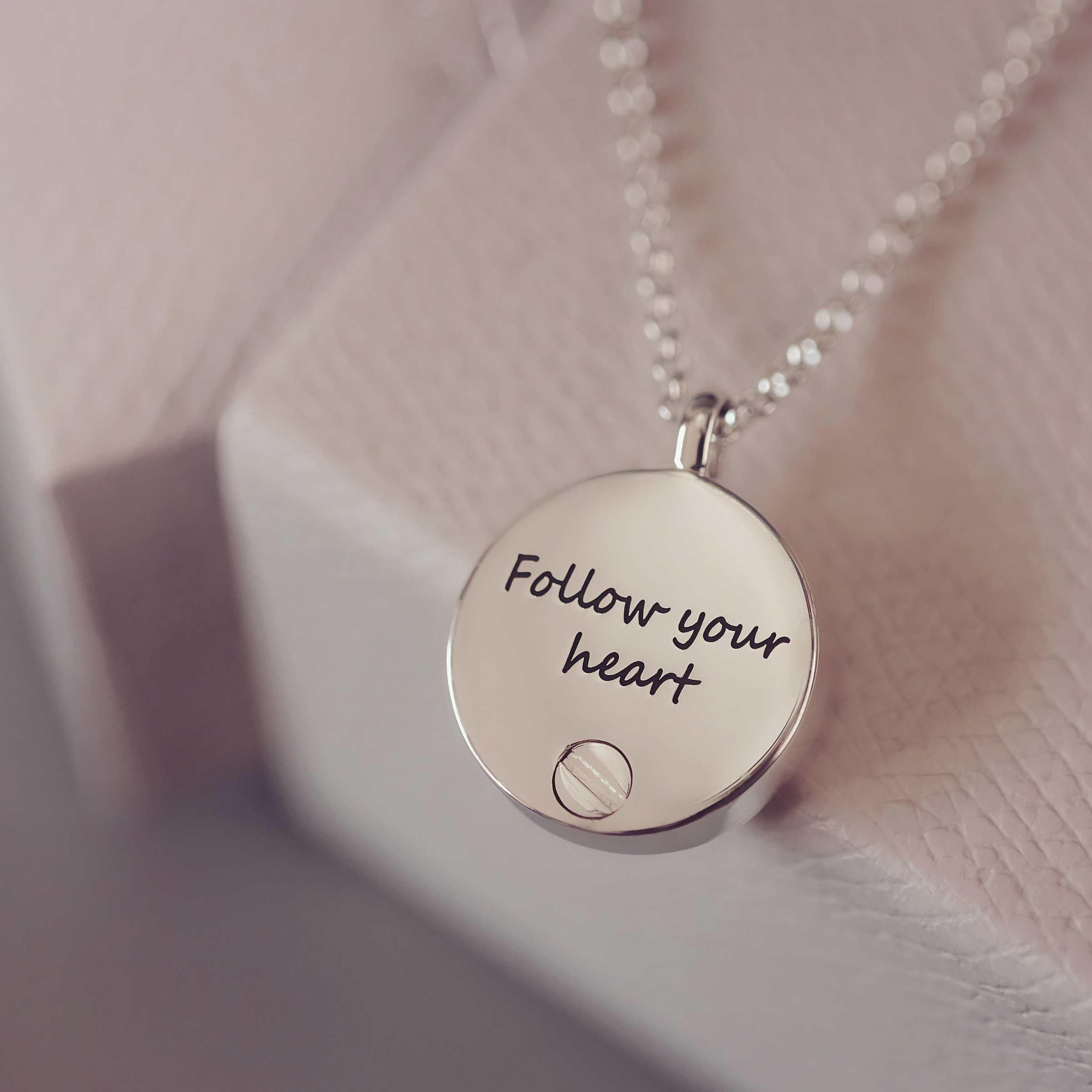 Silver necklace with a round pendant that says 'Follow your heart' on a soft pink background.