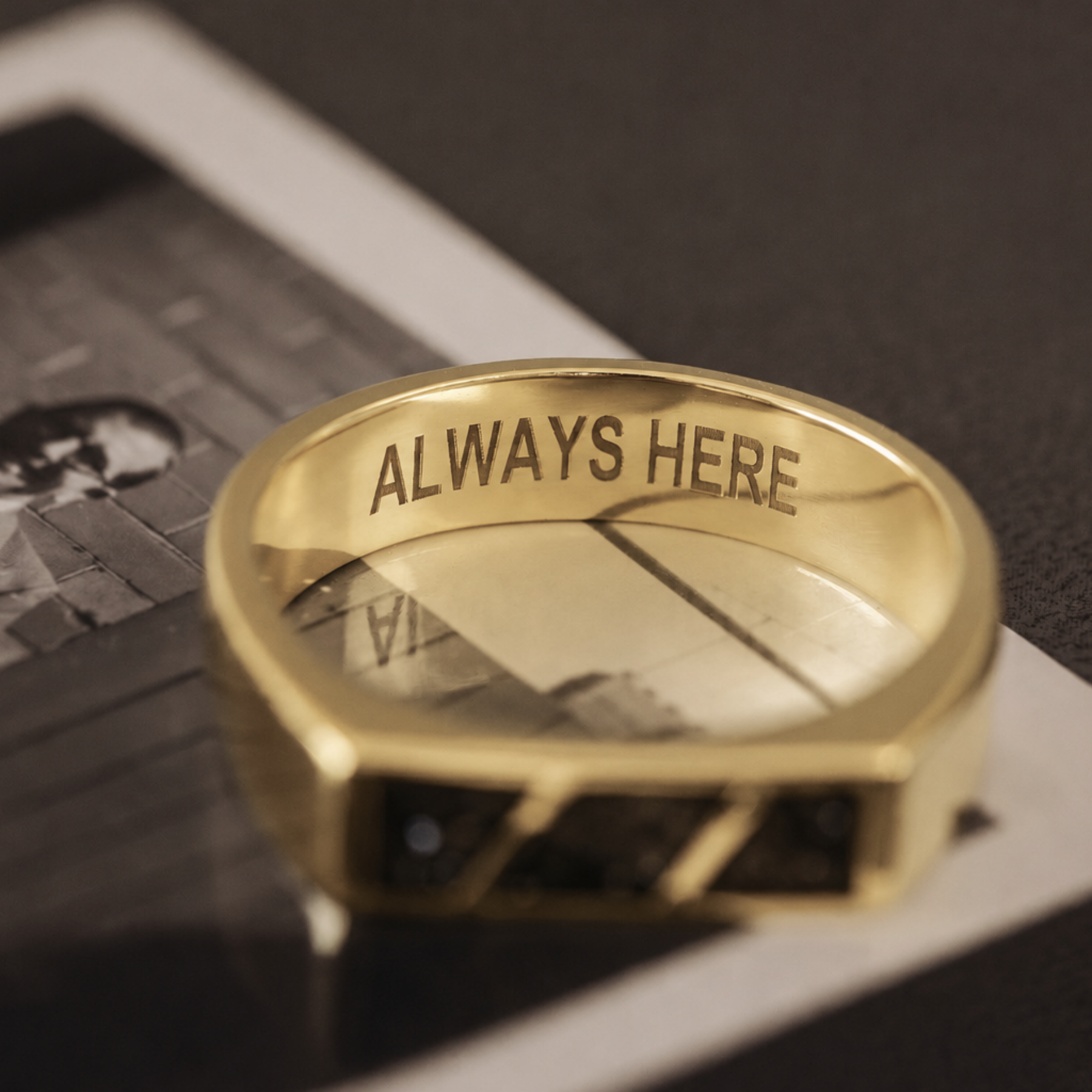 A gold-toned ring engraved with the words 'ALWAYS HERE' on the inside band, resting on a black-and-white printed card over a dark surface.