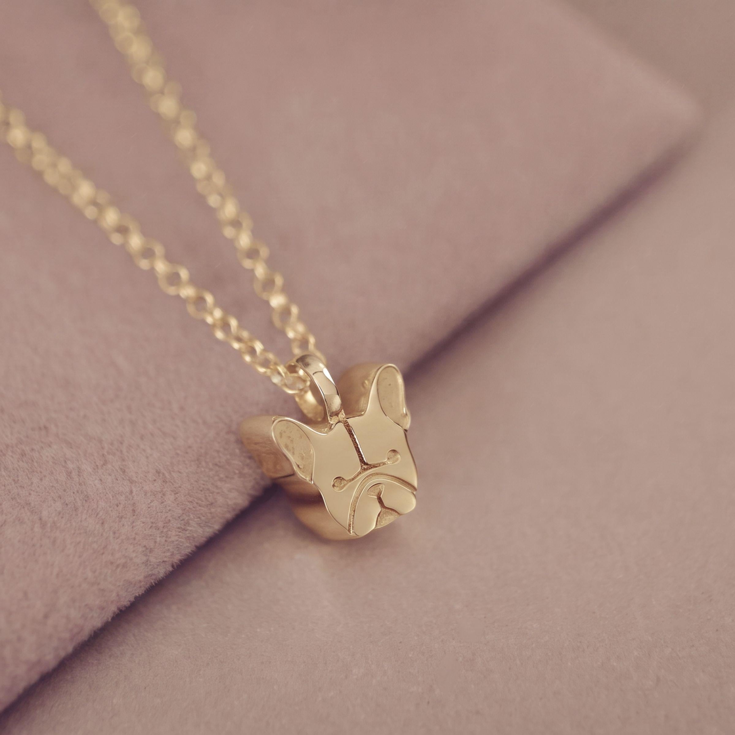 A gold French Bulldog pendant necklace on a delicate gold chain, resting on a soft, dusty pink textured surface.