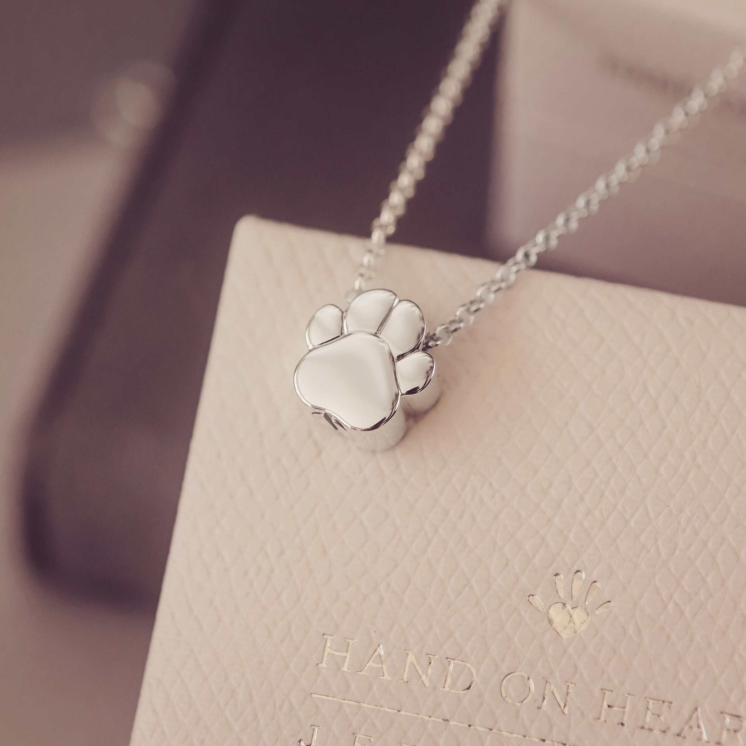 Silver paw print necklace on a beige stand with 'Hand on Heart' branding.