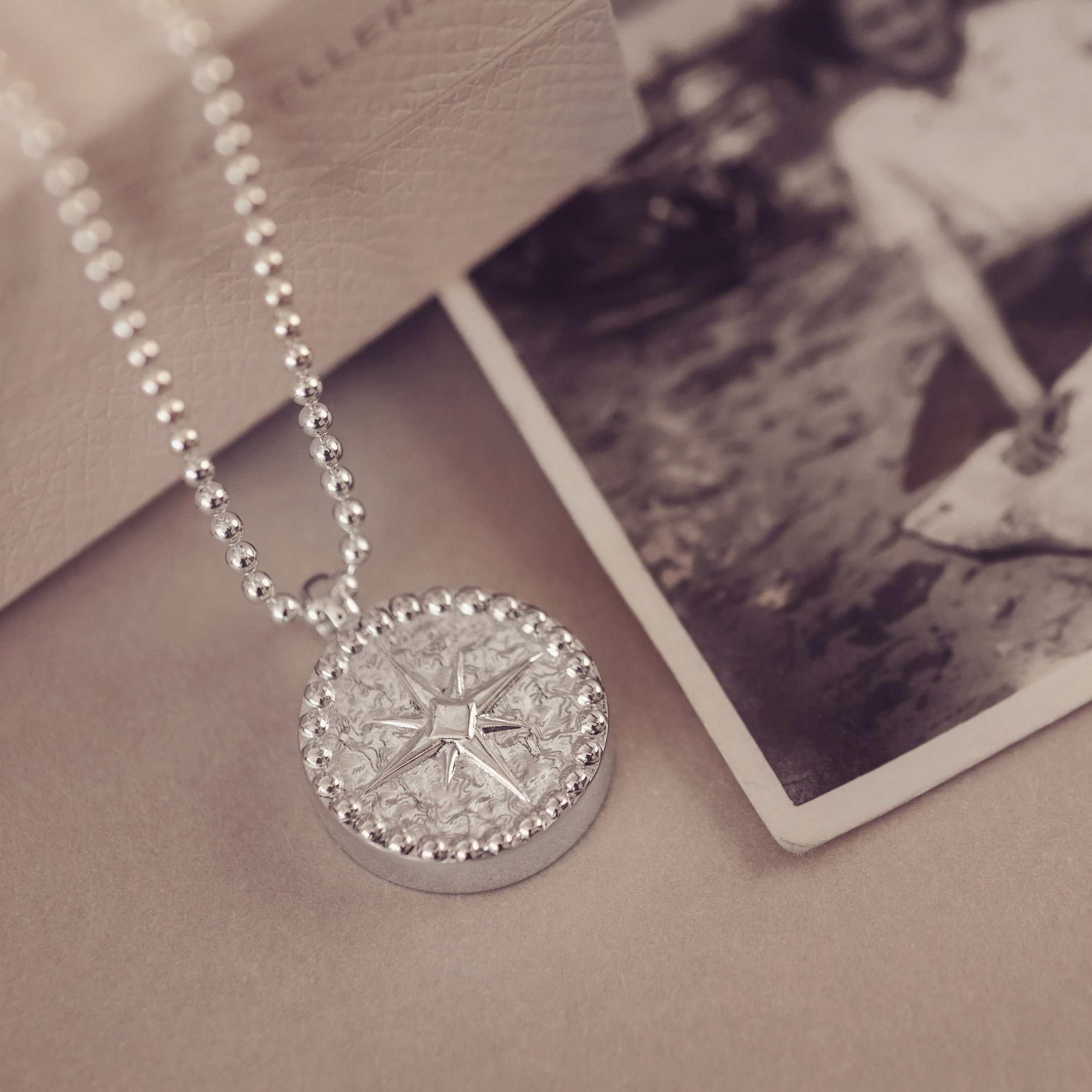 Silver necklace with a round compass pendant on a textured surface next to a vintage photograph.