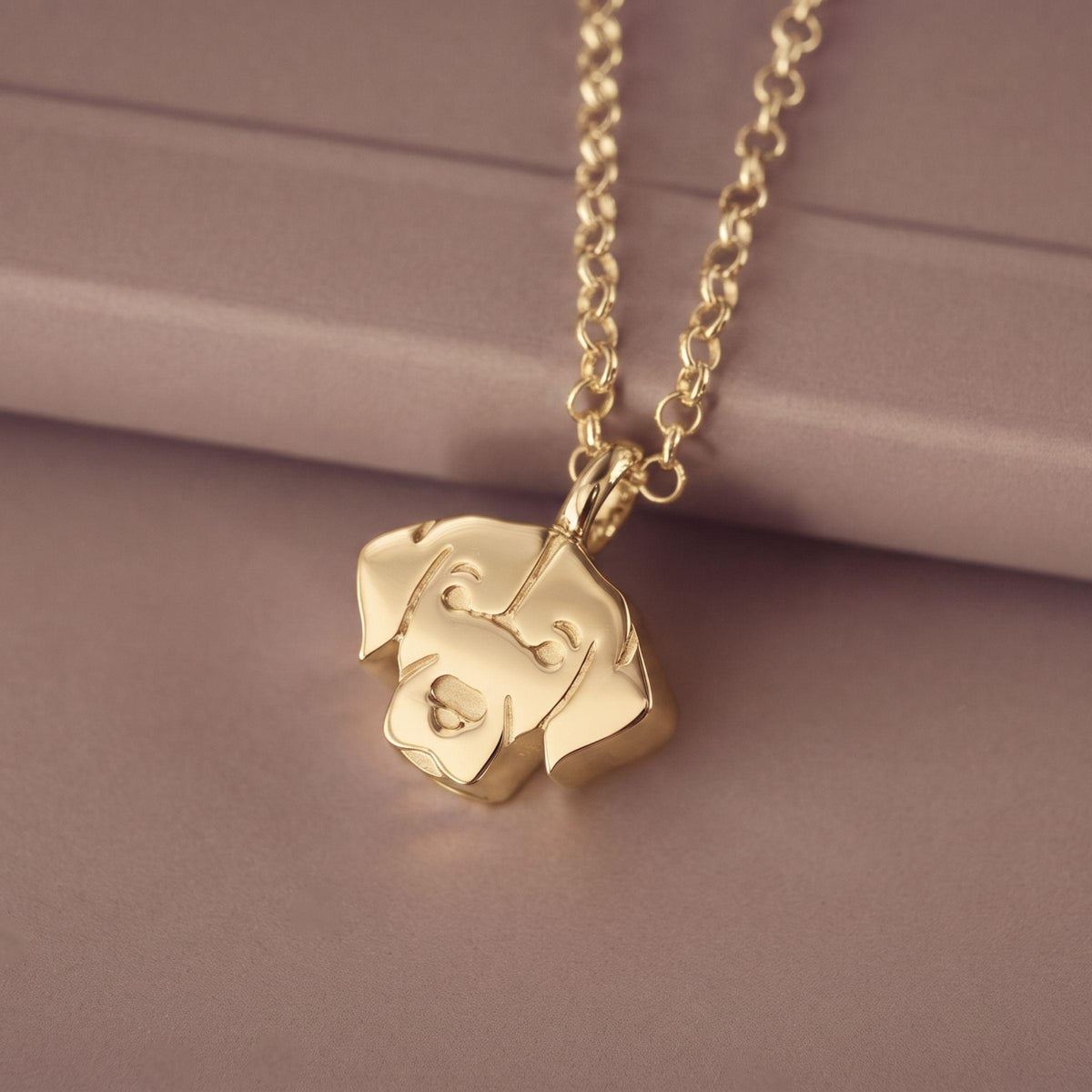 A gold pendant necklace featuring a stylized labrador head outline on a matching chain, displayed on a soft mauve-colored surface.