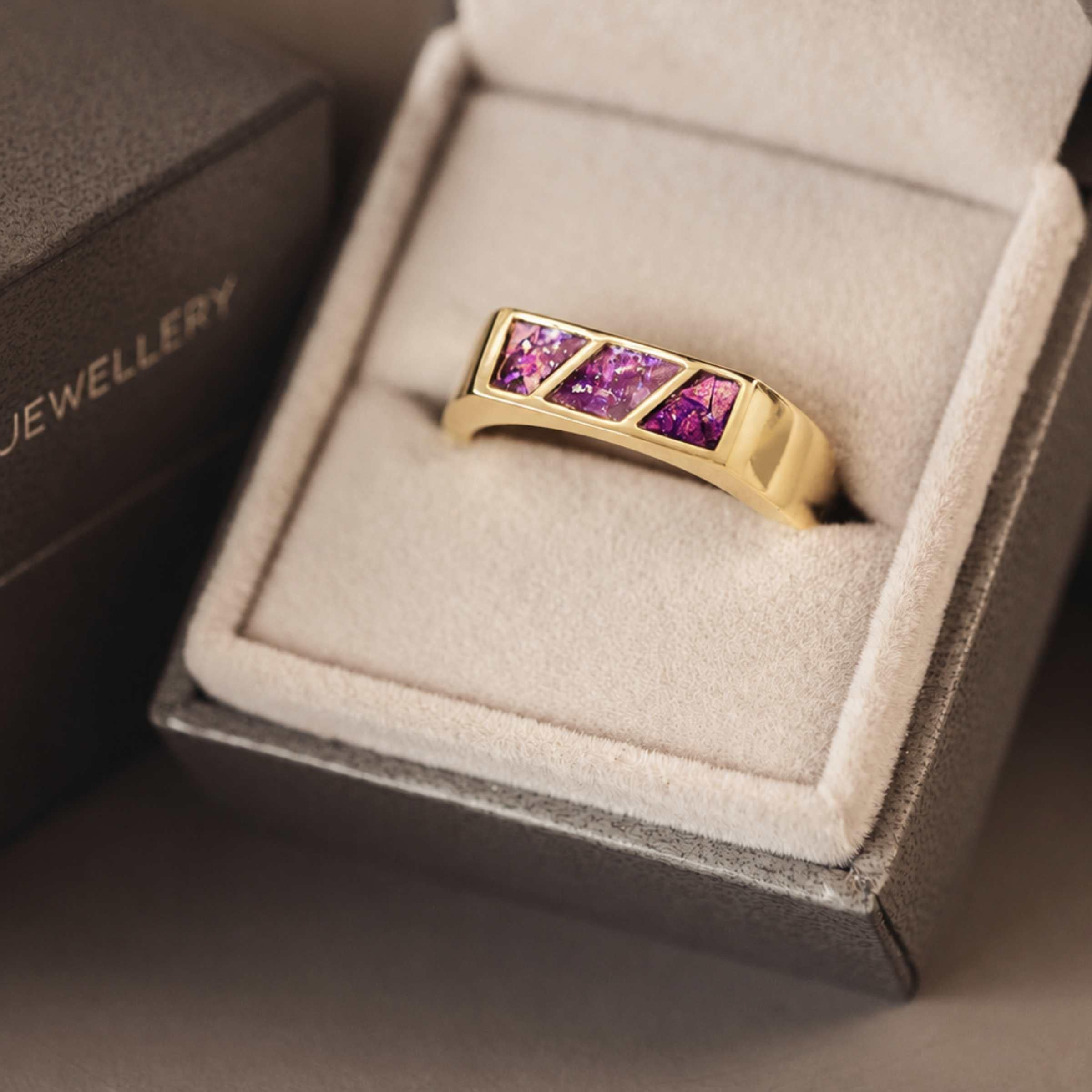 A gold ring with 3 sections of purple resin across the top sitting in an open jewellery box.