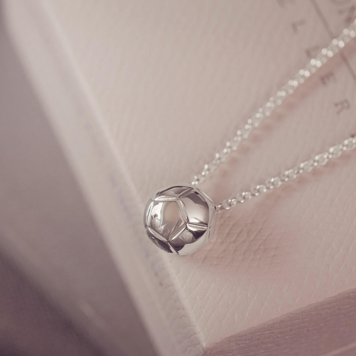 A close-up of a silver pendant necklace with a football, geometric ball design on a delicate chain, resting on a textured light pink surface.
