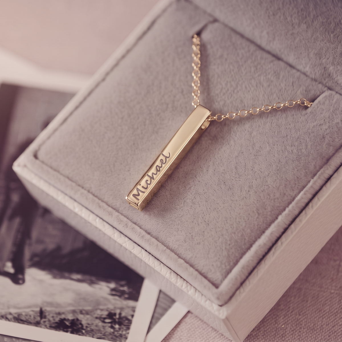 A gold bar necklace engraved with the name 'Michael' in script, resting on a gray velvet jewelry box.