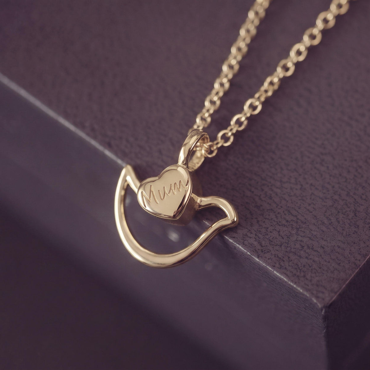 A gold necklace with a Dove & heart-shaped pendant engraved with 'Mum', resting on a dark textured surface.