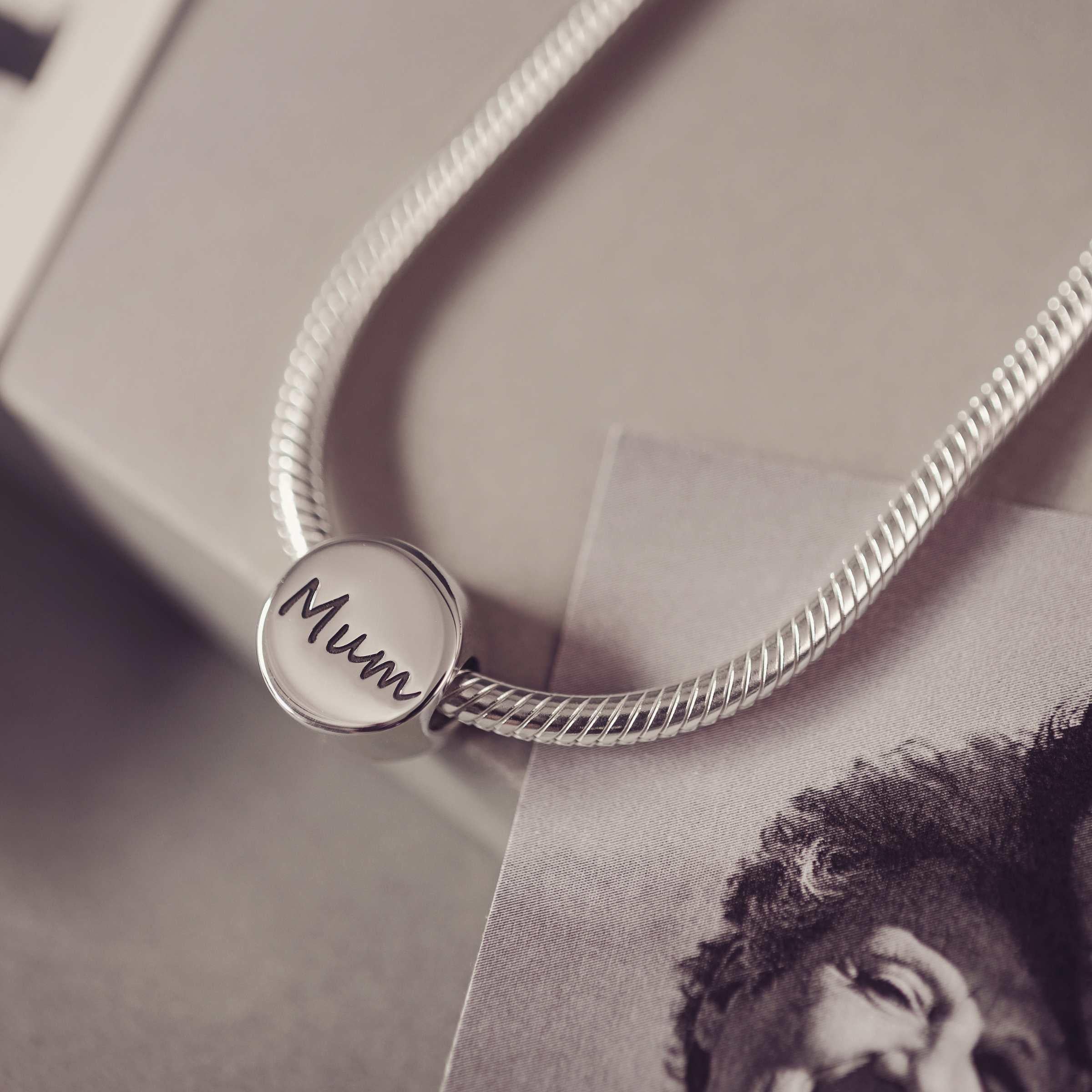 Round bracelet charm engraved with 'Mum' sitting on a thick snake chain. Grey background along side a black and white photograph. 