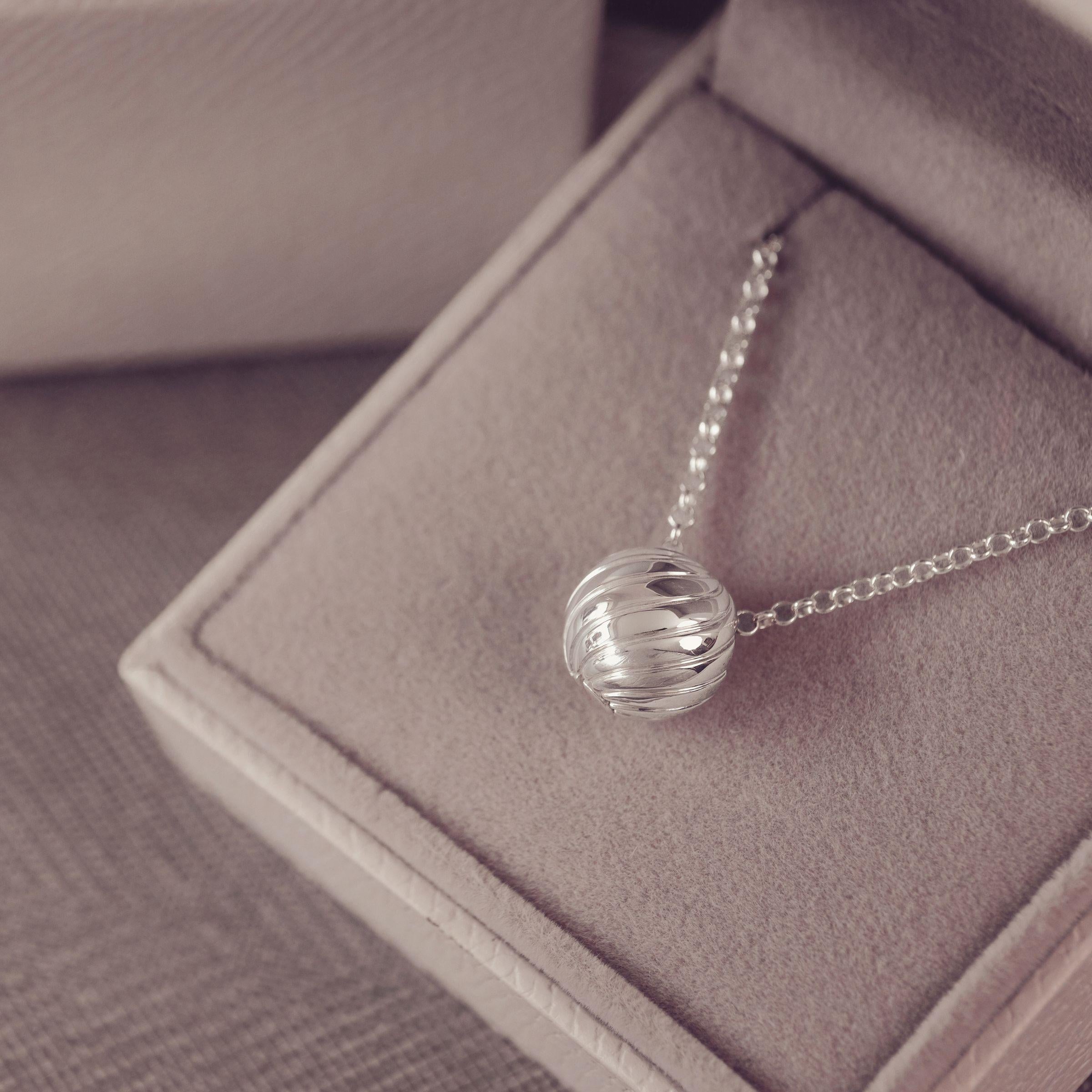 A silver beaded chain necklace with a wave spherical pendant, displayed inside a soft taupe jewelry box.