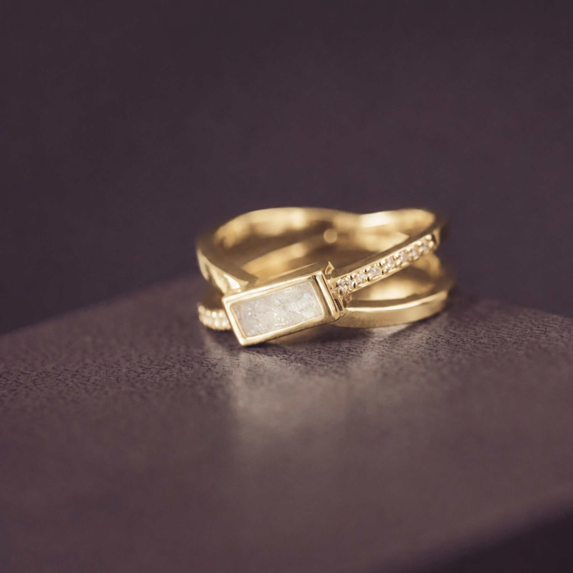 gold criss cross ring with a white rectangle ash stone and gemstones on a dark background