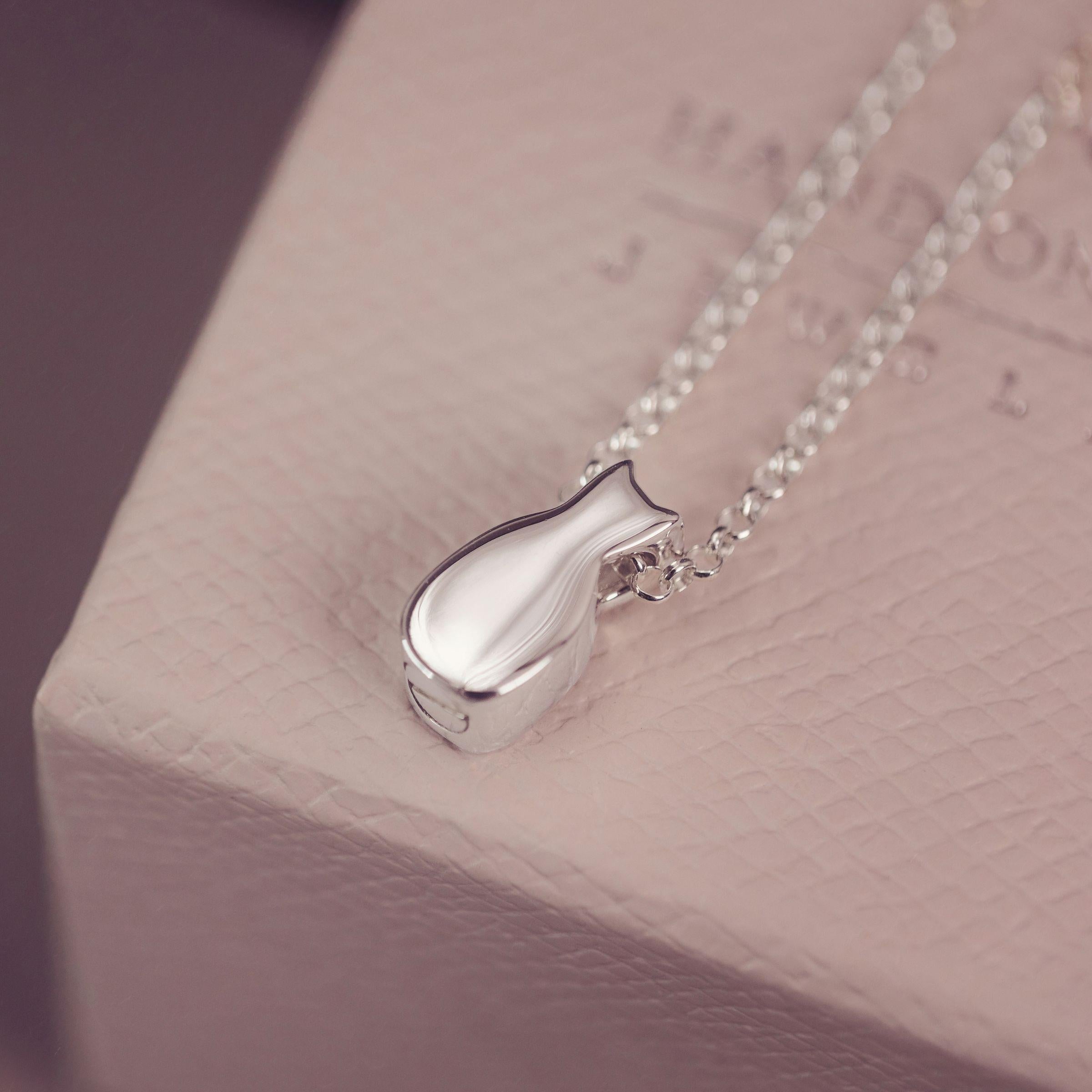 A silver pendant necklace with a cat-shaped charm resting on a textured light pink jewelry box.