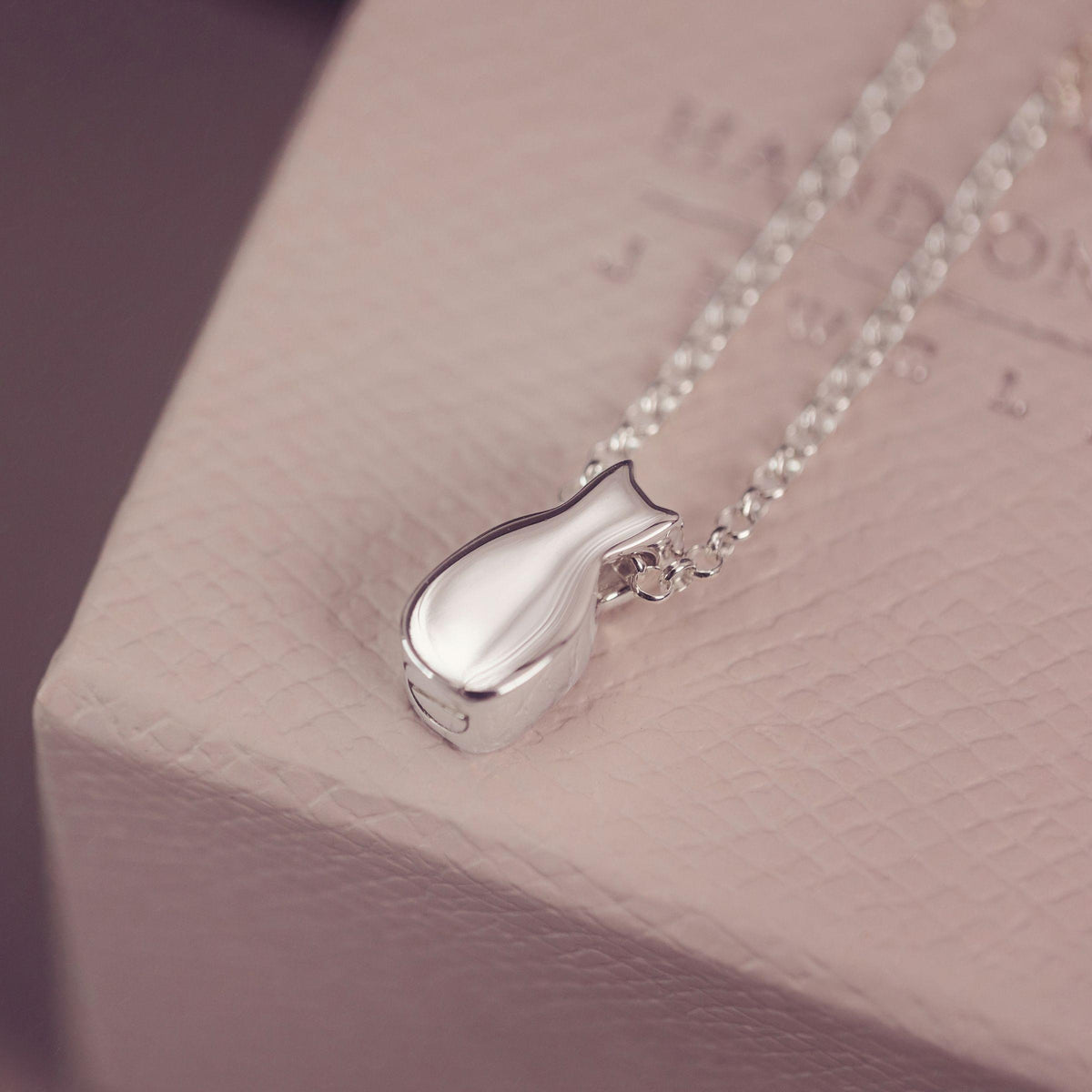 A silver pendant necklace with a cat-shaped charm resting on a textured light pink jewelry box.