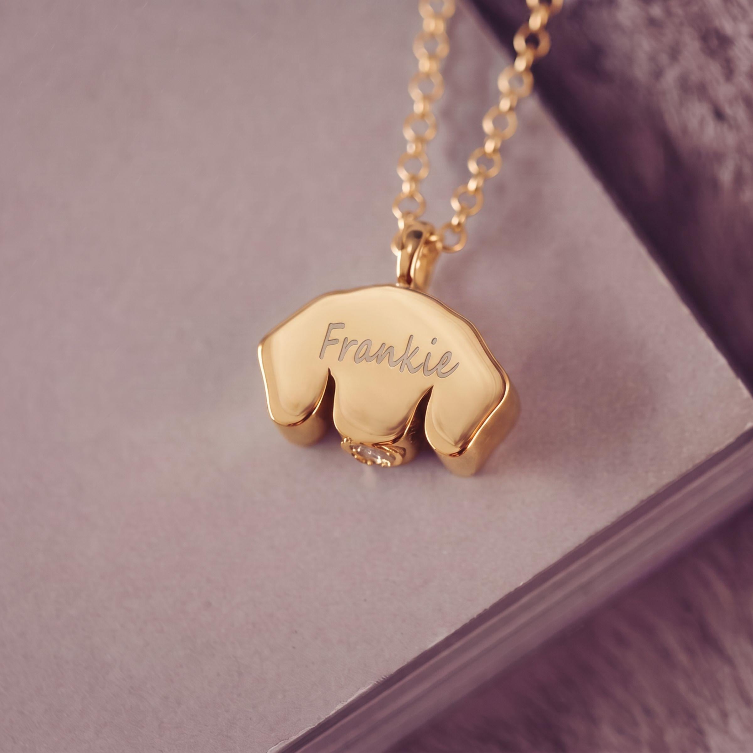 A gold necklace with a small pendant shaped like a Dachshund, engraved with the name 'Frankie', resting on a textured purple surface.