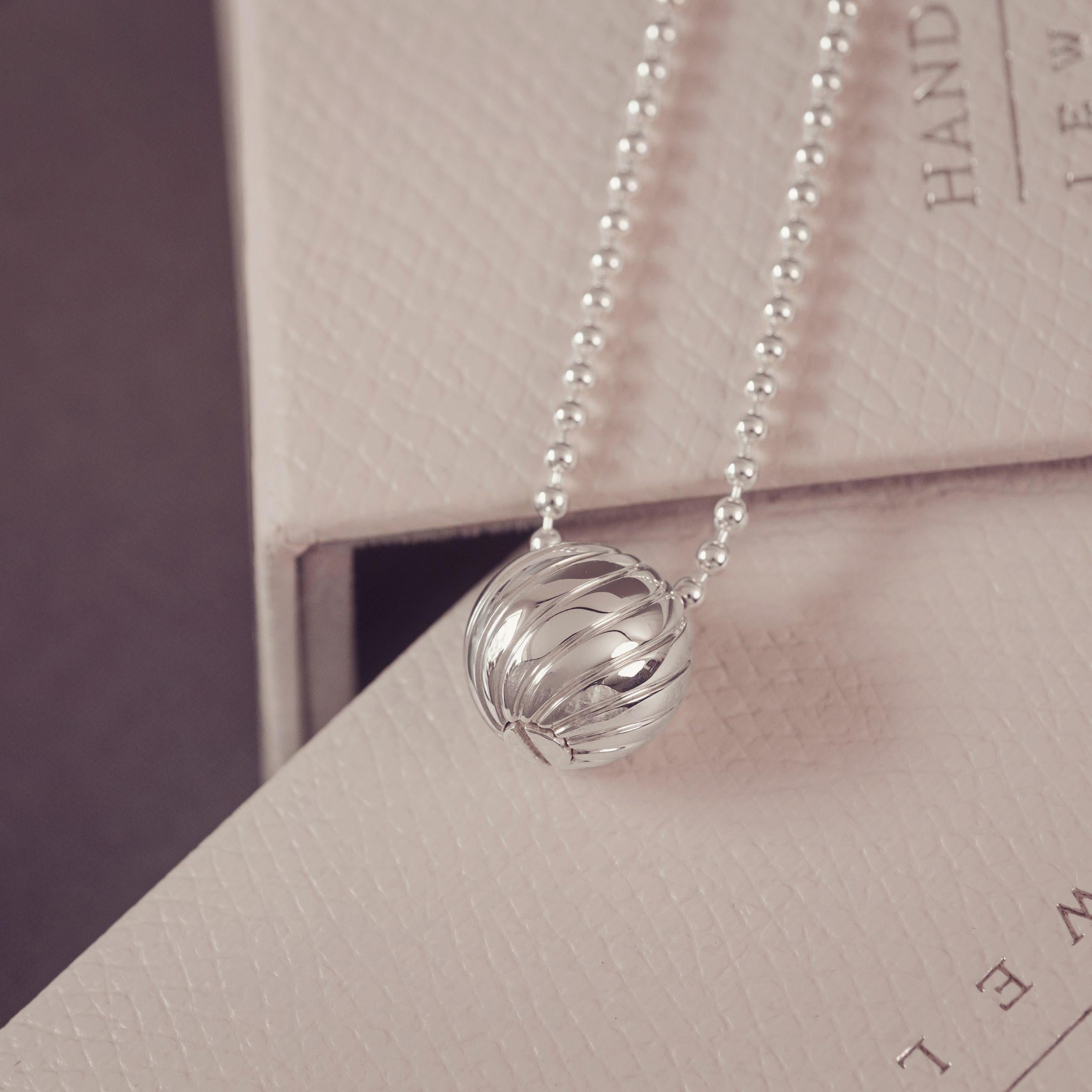 A silver beaded necklace with a textured, ribbed spherical pendant, resting on a light beige textured box.