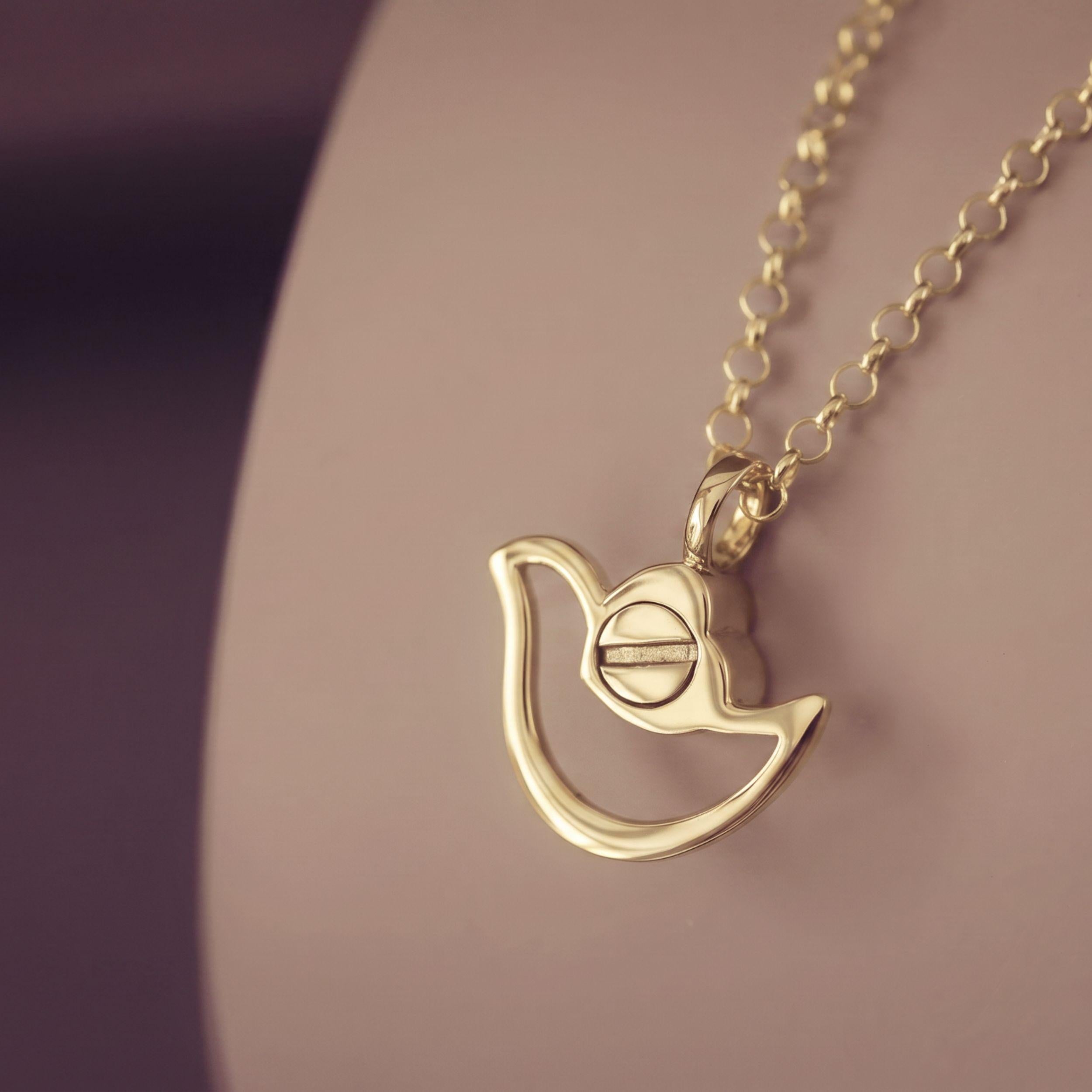 A gold pendant necklace featuring a stylized dove shape with a central screw-like detail, shown resting on a soft beige display surface against a dark purple background.
