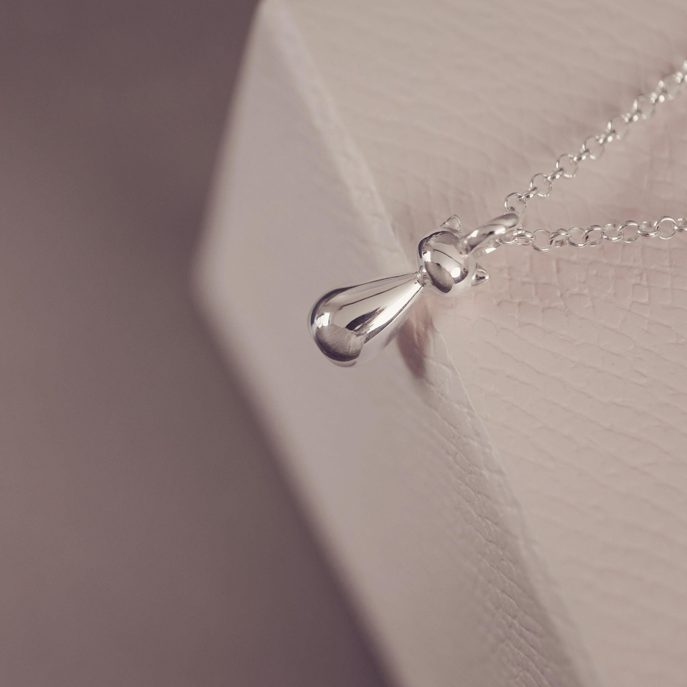 A close-up of a silver stylised cat pendant necklace on a delicate chain, resting against a textured white display block.