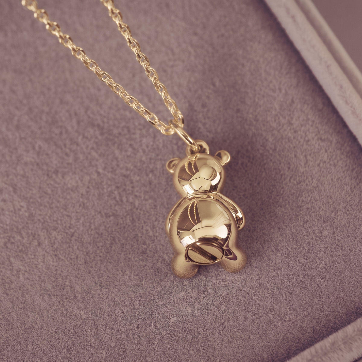 A gold teddy bear pendant necklace resting on a textured light jewellery box, showing the back of the bear and a portion of the chain.