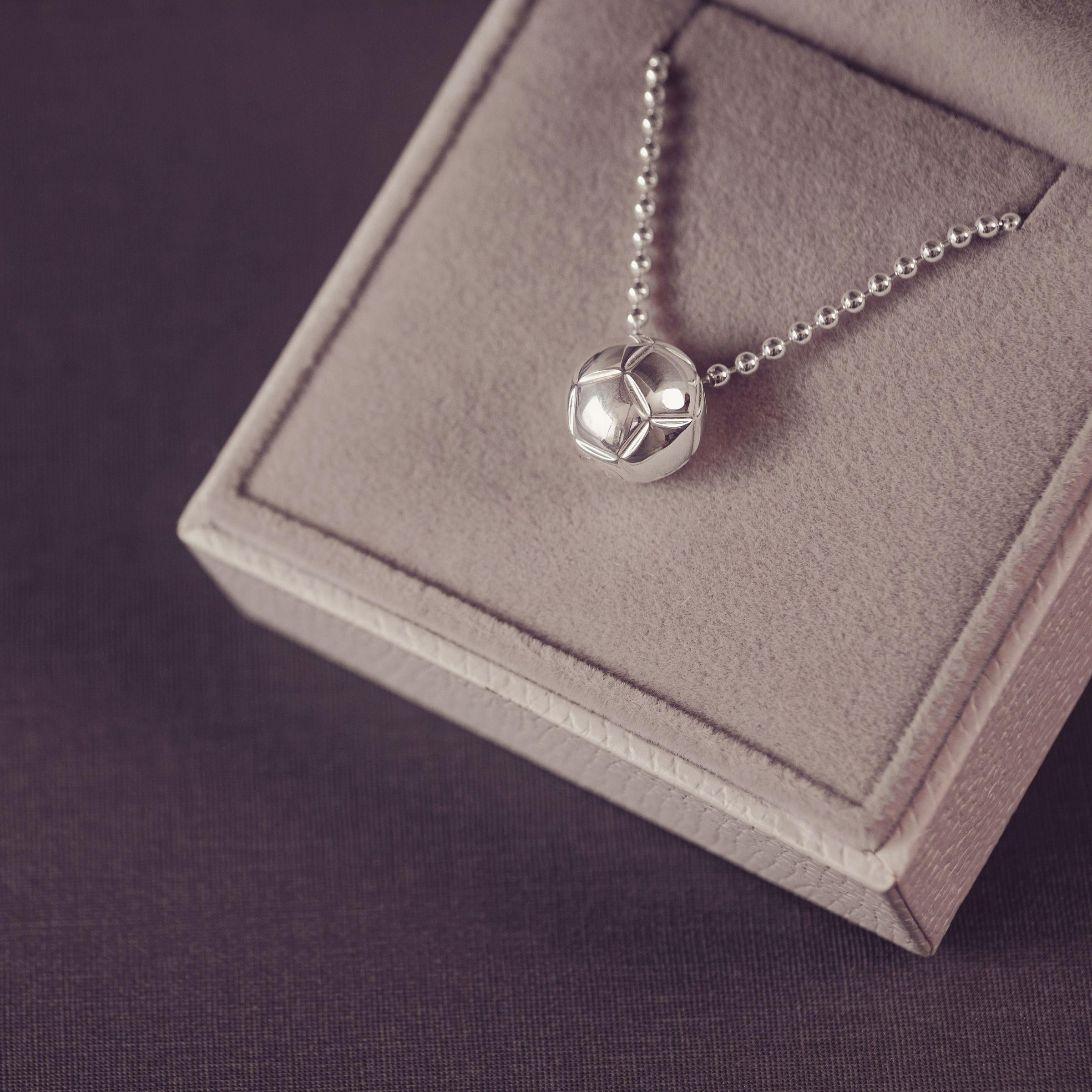 A floating silver football necklace on a beaded chain, displayed on a gray velvet jewelry box against a dark purple background.
