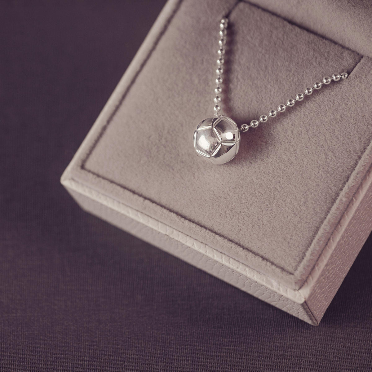 A floating silver football necklace on a beaded chain, displayed on a gray velvet jewelry box against a dark purple background.