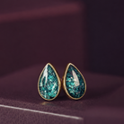 A pair of gold teardrop stud earrings with textured, glittery teal-green resin centers, resting on a dark purple surface.
