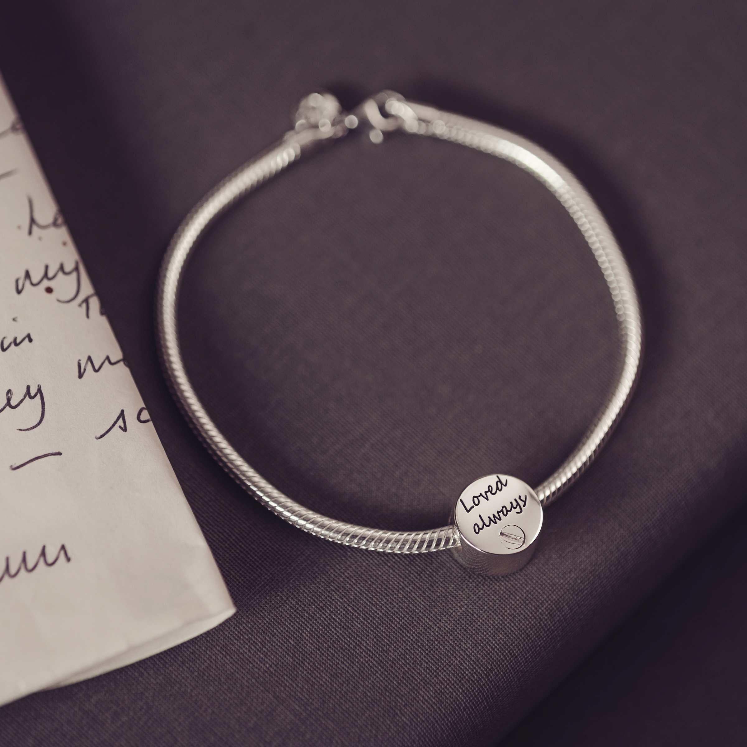 Round bracelet urn charm engraved with 'loved always' sitting on a thick snake chain bracelet.dark background along side a letter. 