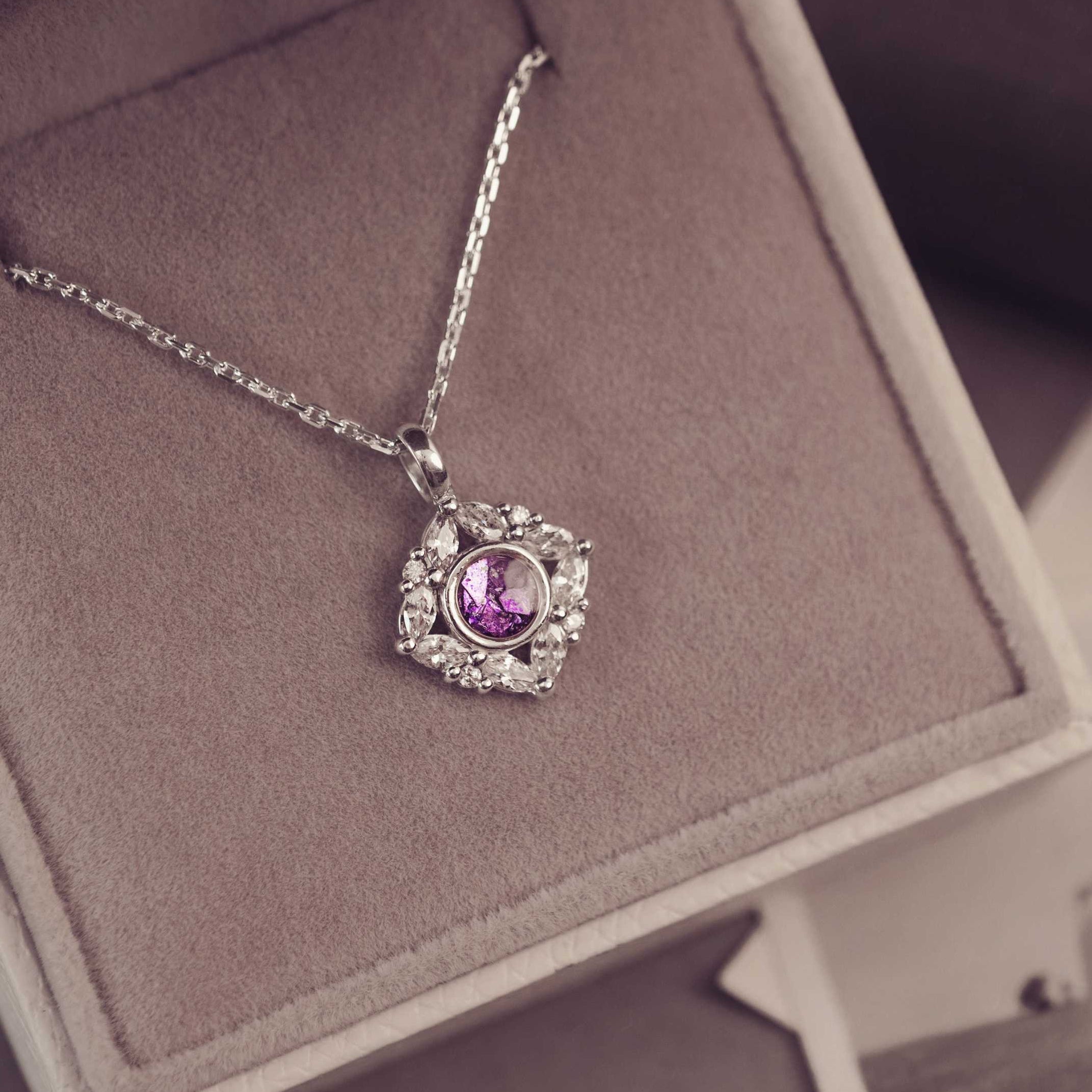 Necklace with a purple gemstone pendant on a pink cushion, with a blurred background of people and nature.