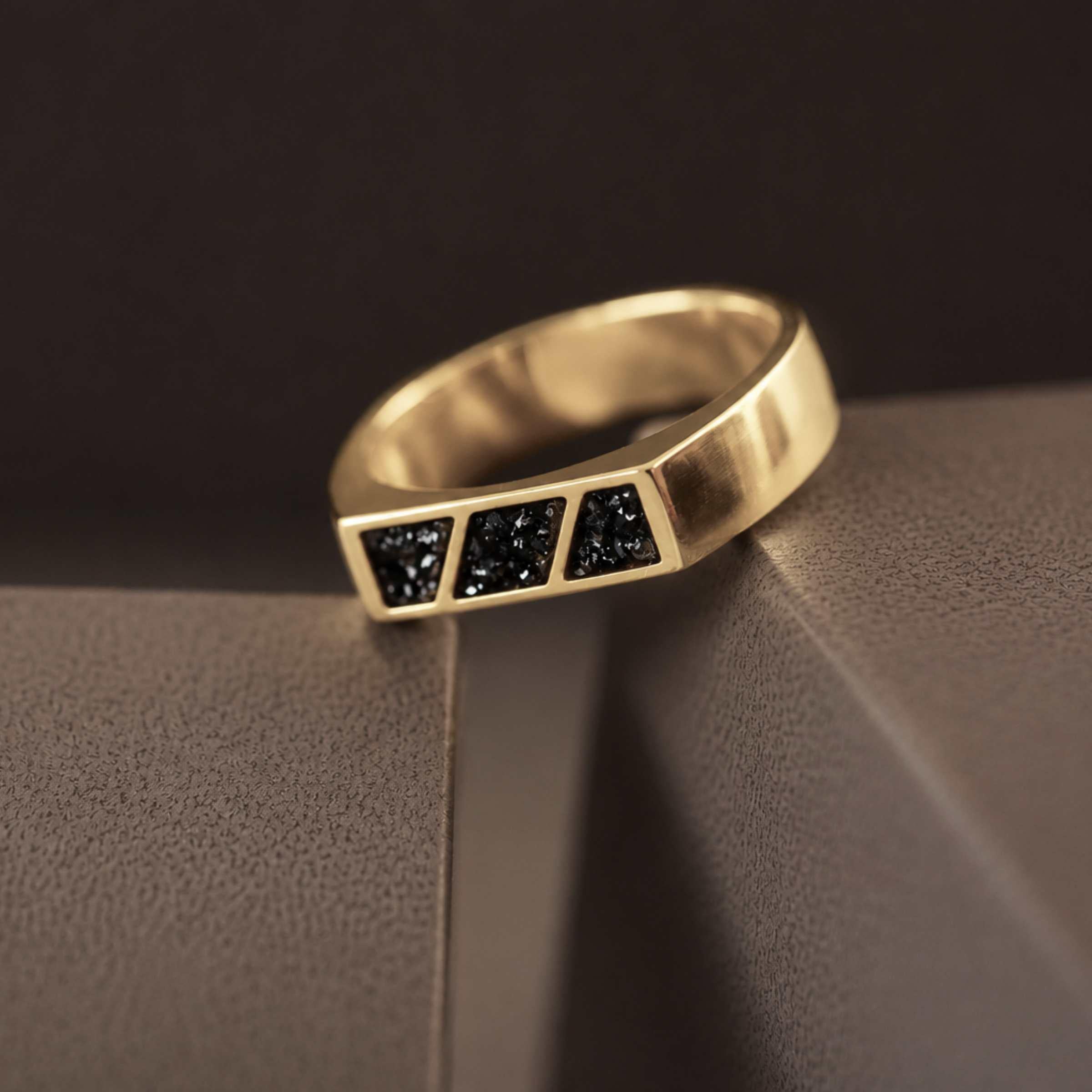 A gold ring with 3 sections of blacked resin across the top. Sitting on a textured dark background