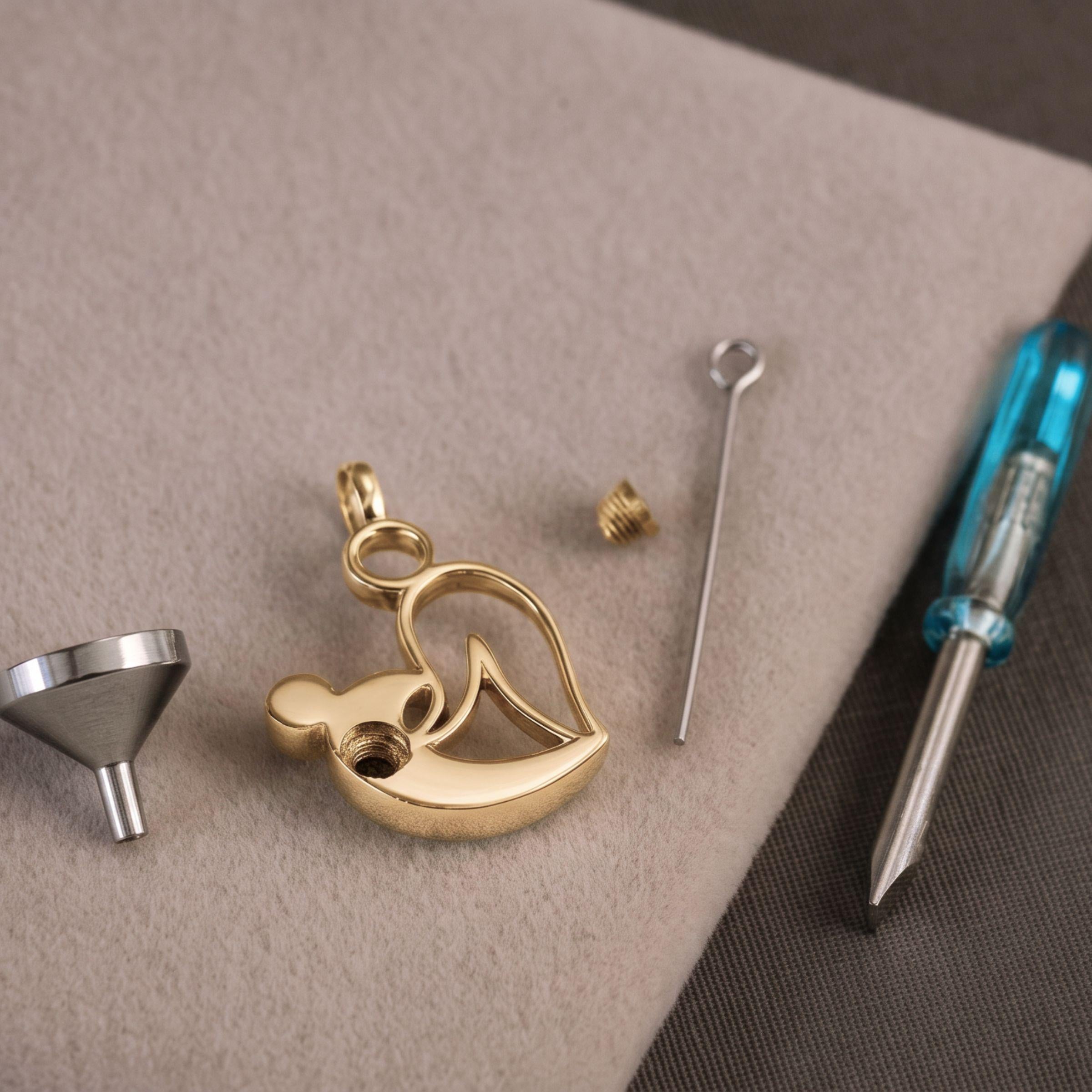 A styalised mother and child pendant charm with a small bail, shown with a silver funnel, a small screw, a pin, and a blue-handled screwdriver on a beige textured surface.