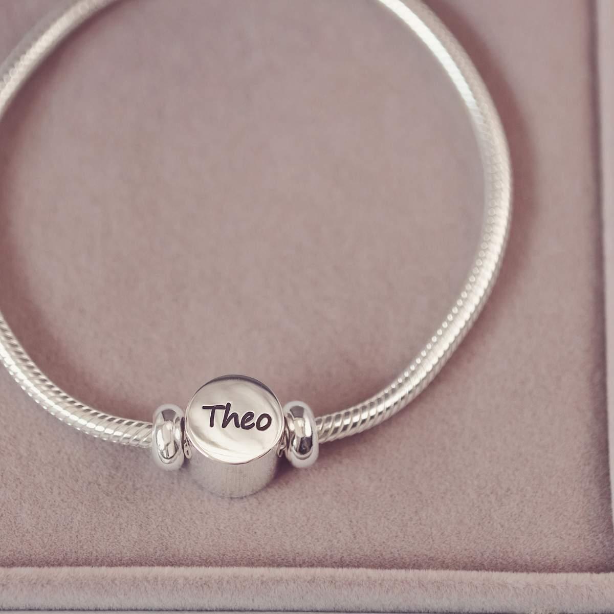 Silver bracelet with a round charm engraved with 'Theo' on a pink background