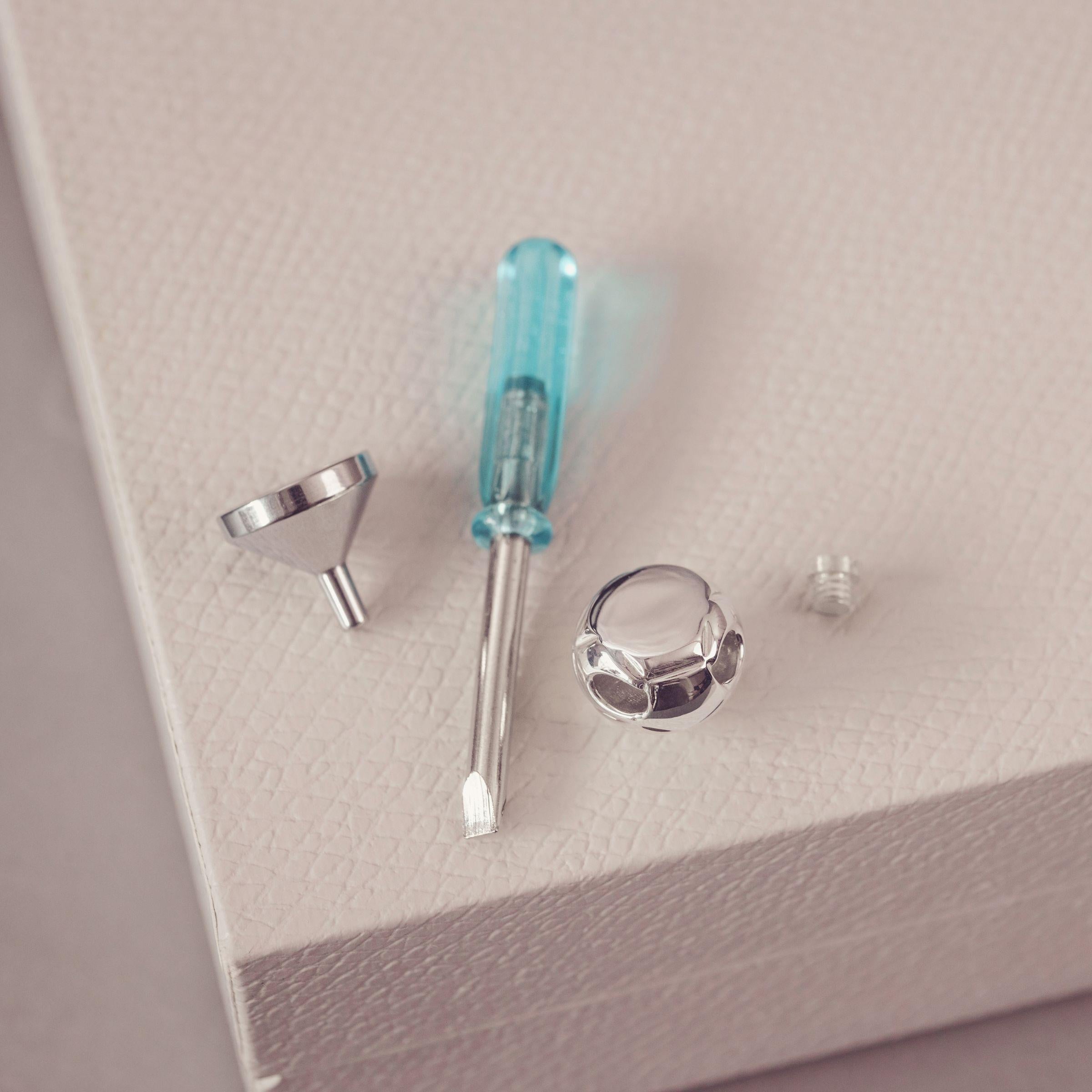 Jewellery maker's tools and components, including a small screwdriver with a translucent blue handle, a metal funnel, and a small metal piece, arranged on a textured light beige surface.