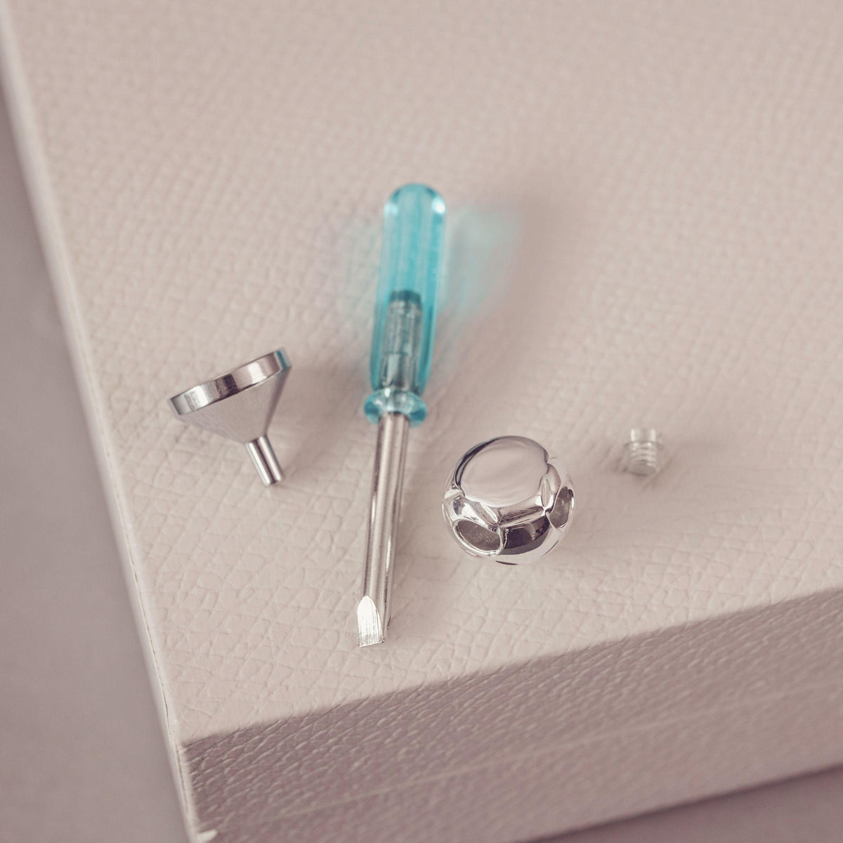 Jewellery maker's tools and components, including a small screwdriver with a translucent blue handle, a metal funnel, and a small metal piece, arranged on a textured light beige surface.