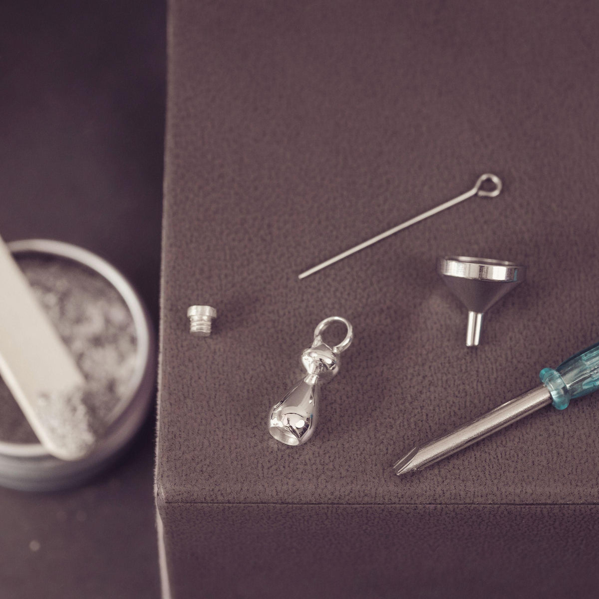 Silver jewelry kit laid out on a dark textured surface, including a small metal screw, a pointed tool, a funnel, and a screwdriver, with a blurred round tin in the background.