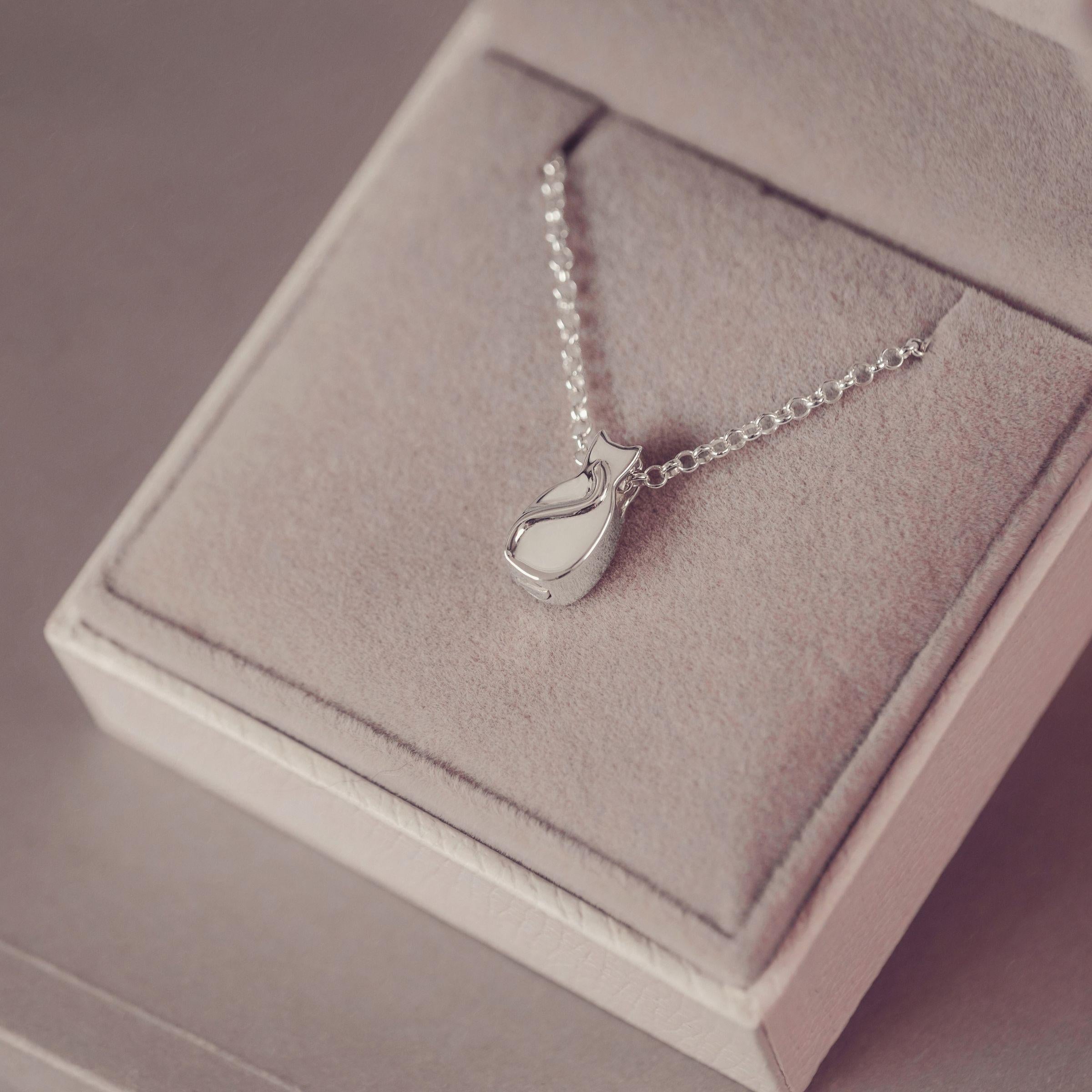 A silver pendant necklace with a small stylized cat silhouette, displayed on a soft pink jewelry box.