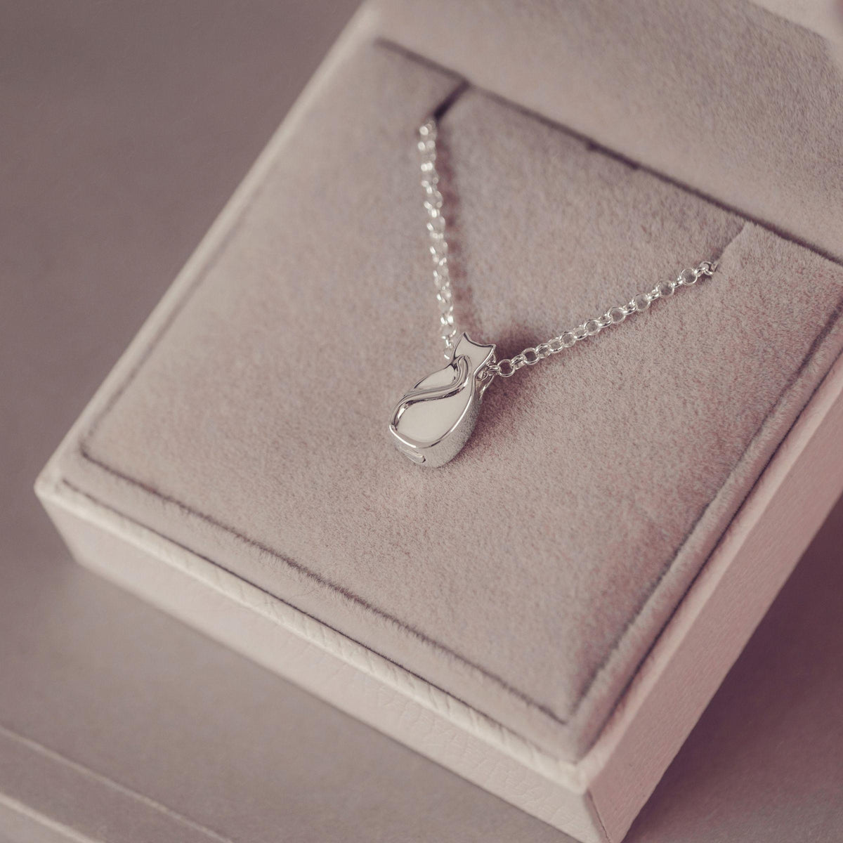 A silver pendant necklace with a small stylized cat silhouette, displayed on a soft pink jewelry box.