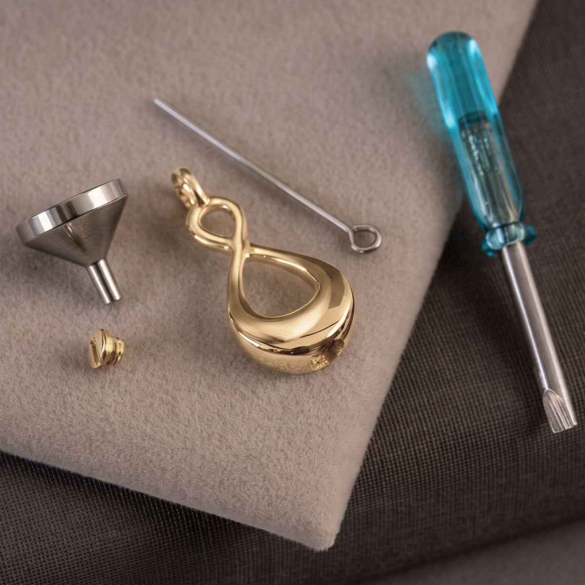 A gold infinity-shaped pendant charm displayed with its silver metal funnel, small gold cap, and a blue-handled screwdriver on a beige textured mat.