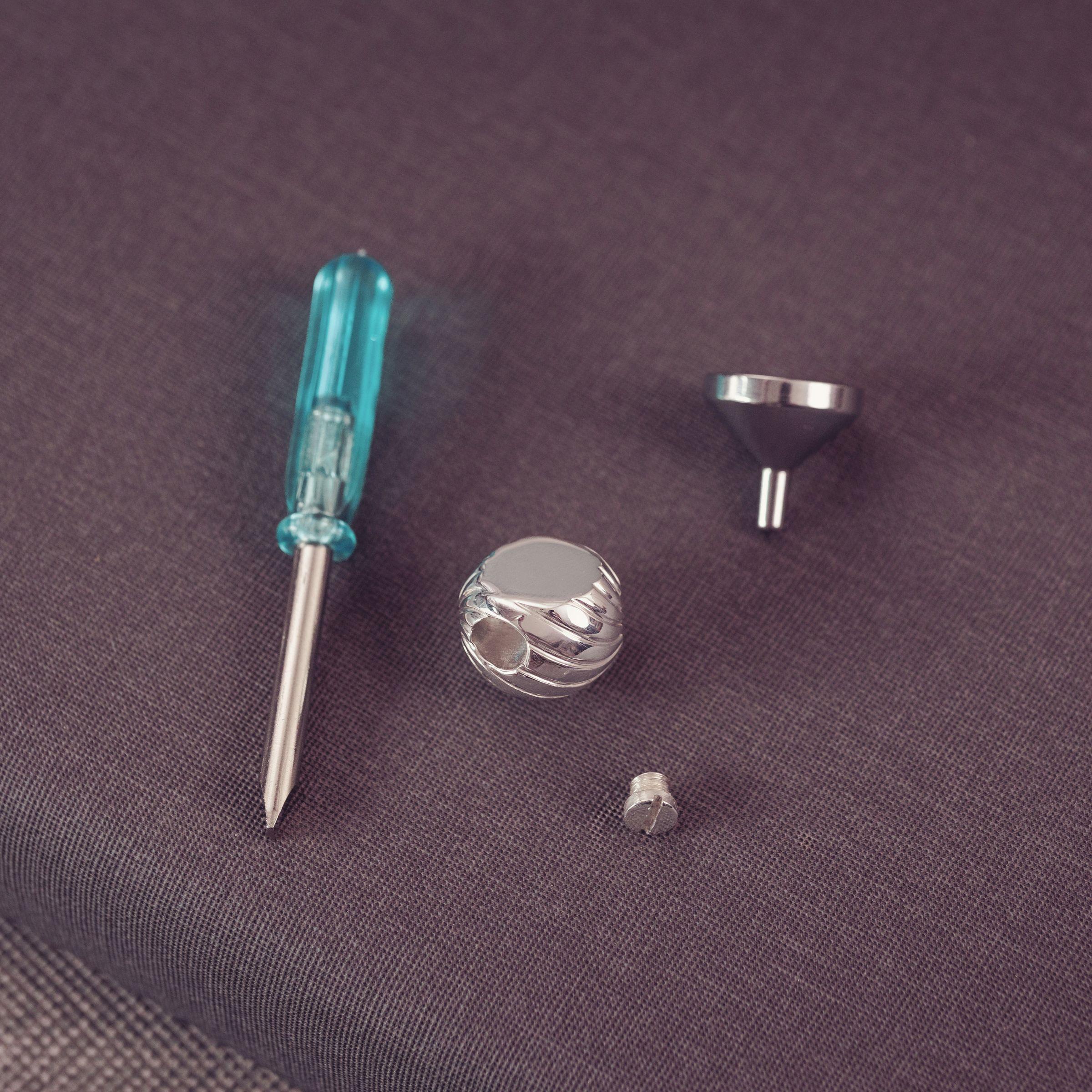 Silver metal jewelry finding set a small screw, and a funnel, alongside a turquoise-handled precision screwdriver, arranged on a textured dark gray fabric background.