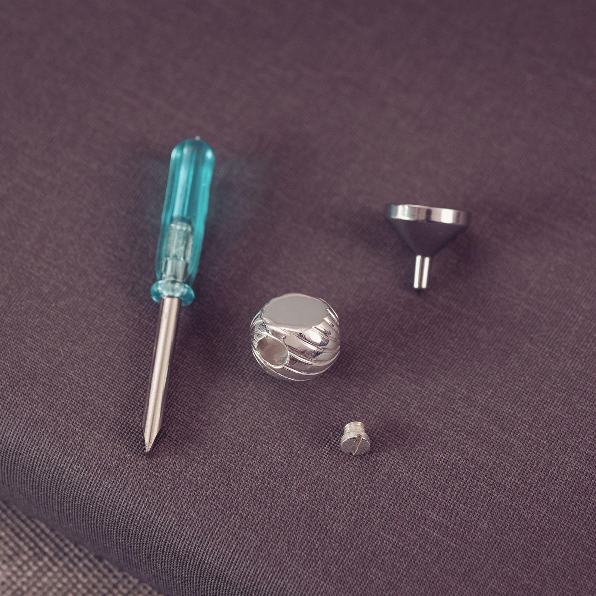 Silver metal jewelry finding set a small screw, and a funnel, alongside a turquoise-handled precision screwdriver, arranged on a textured dark gray fabric background.