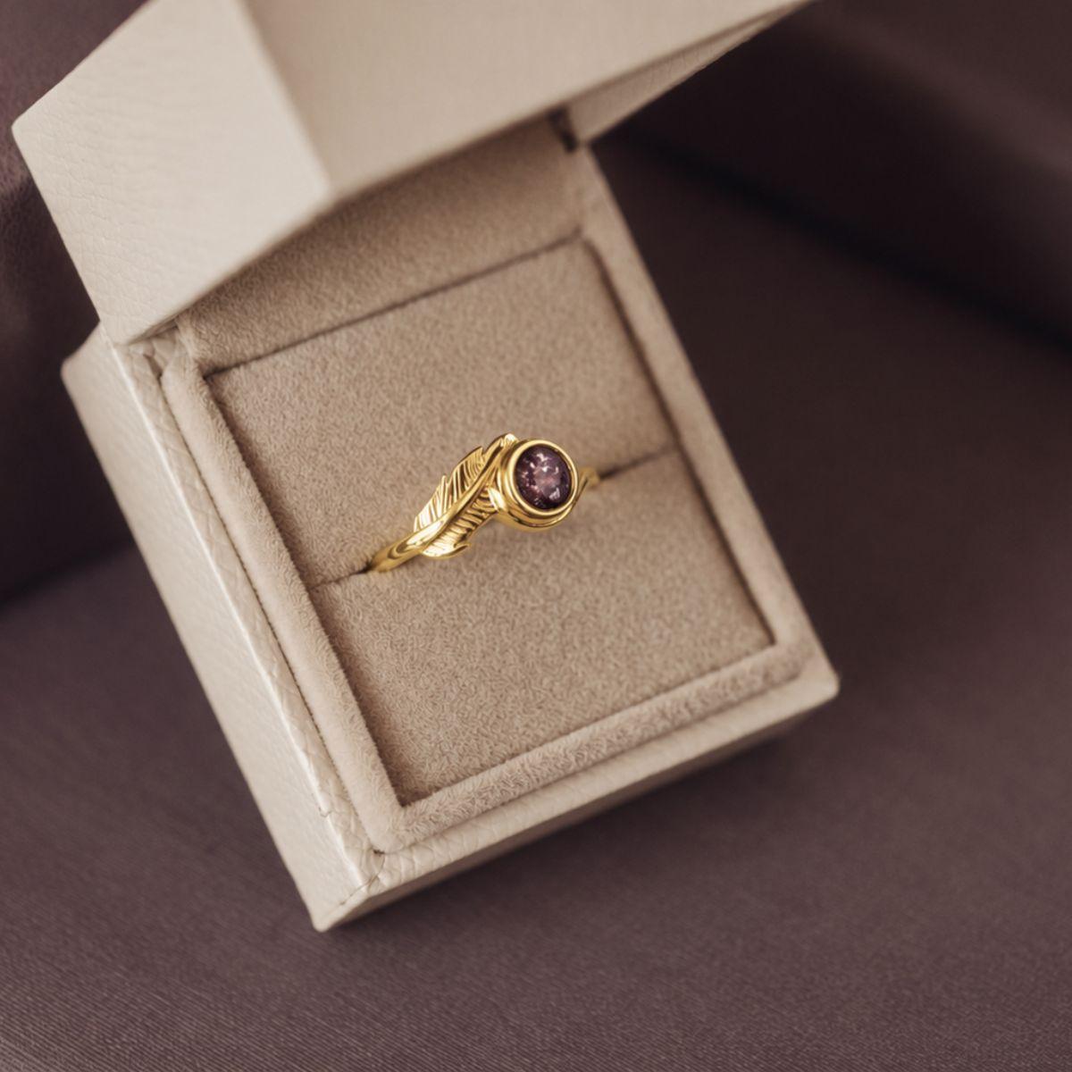 A gold ring with a leaf-shaped band and a round purple gemstone, displayed inside an open beige jewelry box on a dark brown surface.